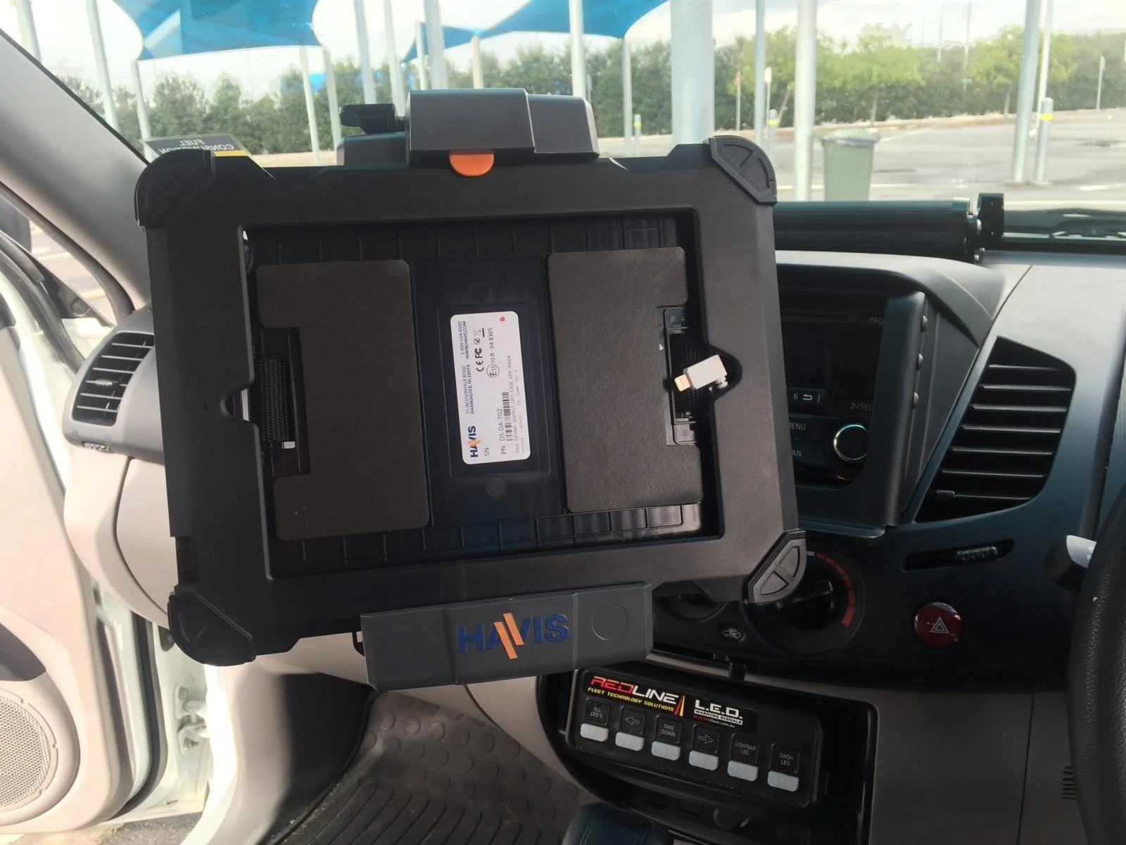 Interior of a vehicle with a mounted tablet on the dashboard, showing a close-up view of the tablet and dashboard controls, with a window revealing an outdoor scene with trees and a blue structure.
