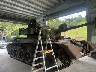 A military tank indoors with a ladder next to it, positioned in a spacious garage or storage facility with open doors revealing green trees and rolling hills outside.