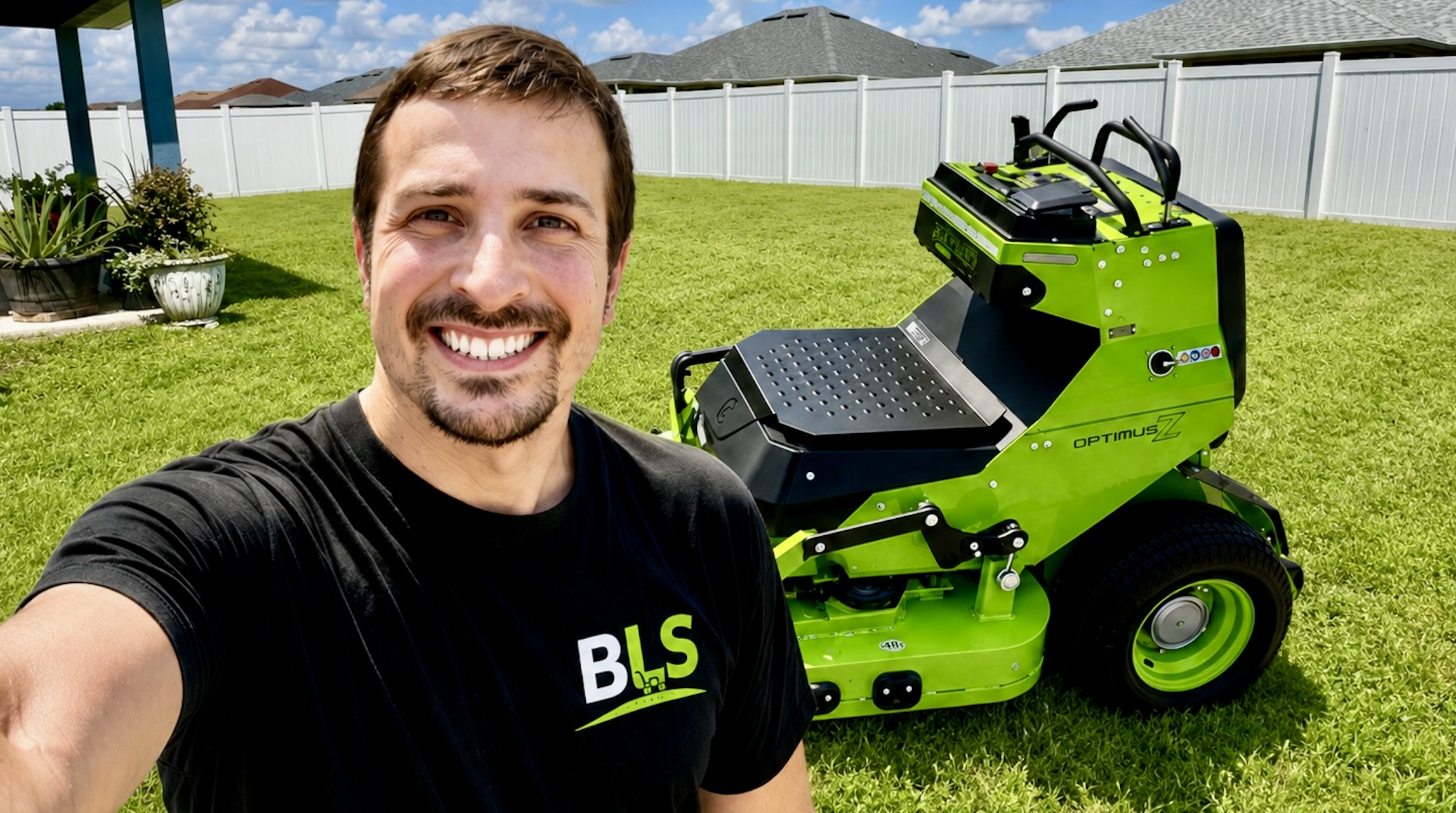 A smiling man taking a selfie in a backyard with green grass, white fence, and houses in the background. Next to him is a bright green lawnmower with the brand name 'Optimus Z' on it.