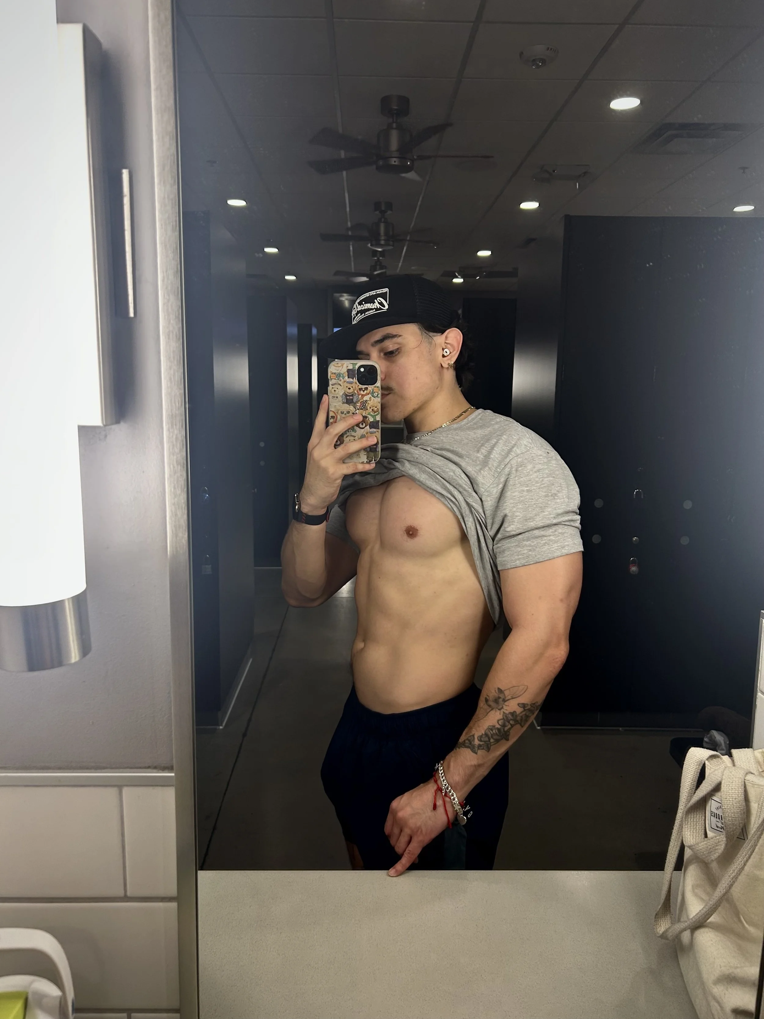 A young man taking a mirror selfie in a bathroom, lifting his gray t-shirt to show his toned abdomen, wearing a black cap, earrings, a gold chain, and dark shorts, with a tattoo on his right arm.