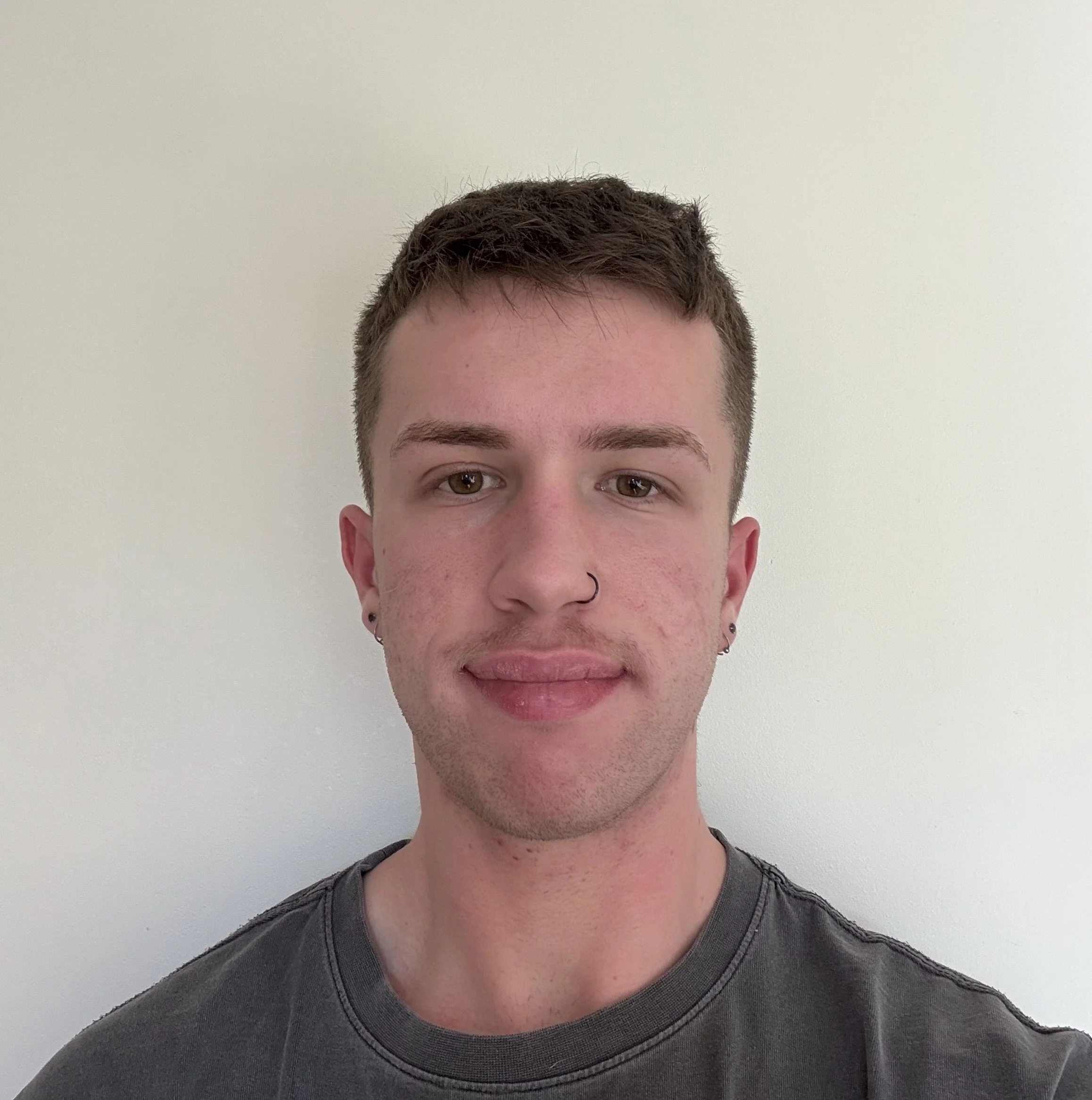 Partial view of a young man with short brown hair and brown eyes, wearing a dark gray shirt, against a plain light-colored wall.