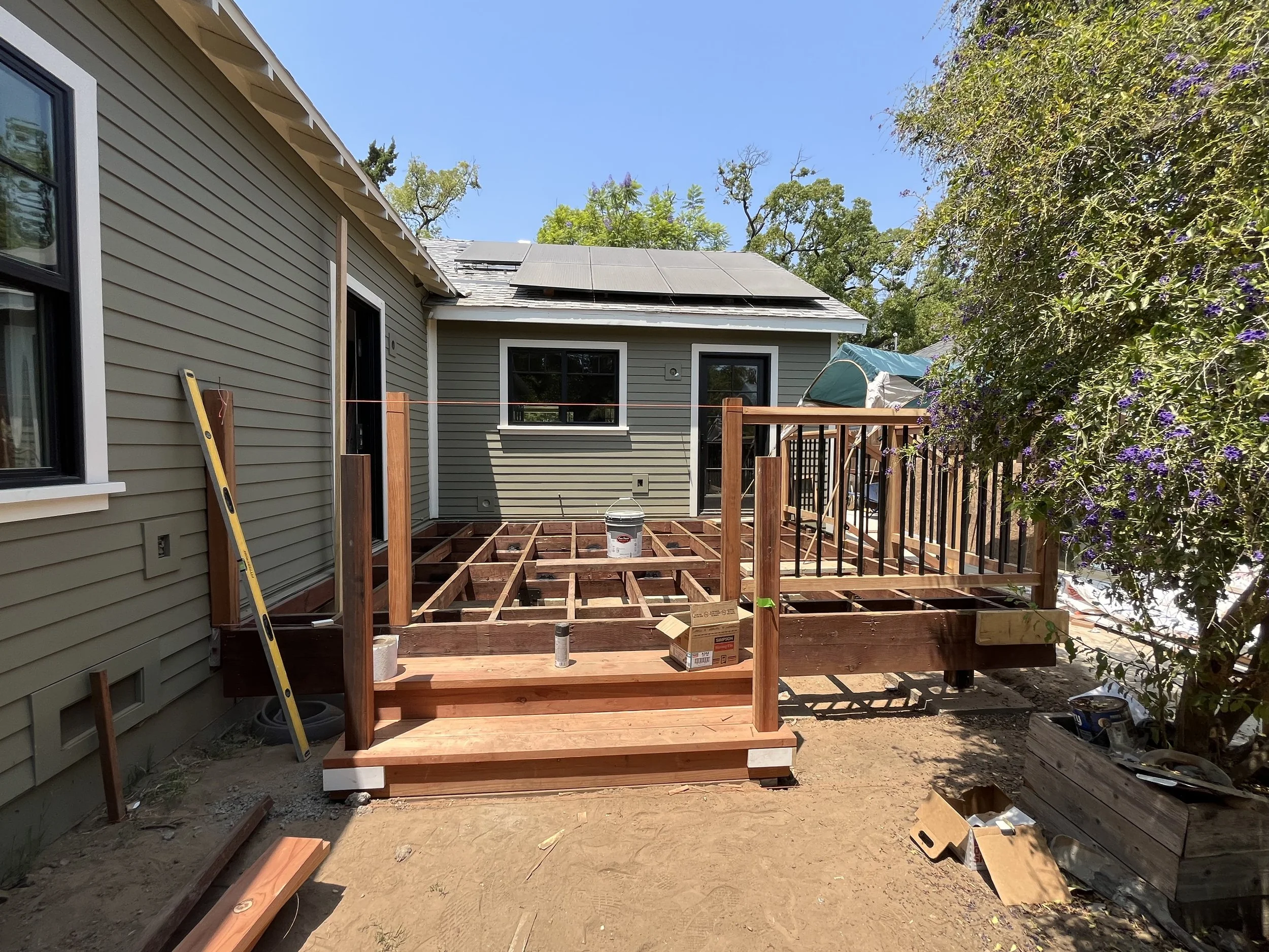 Backyard deck framing under construction beside a gray home with solar panels