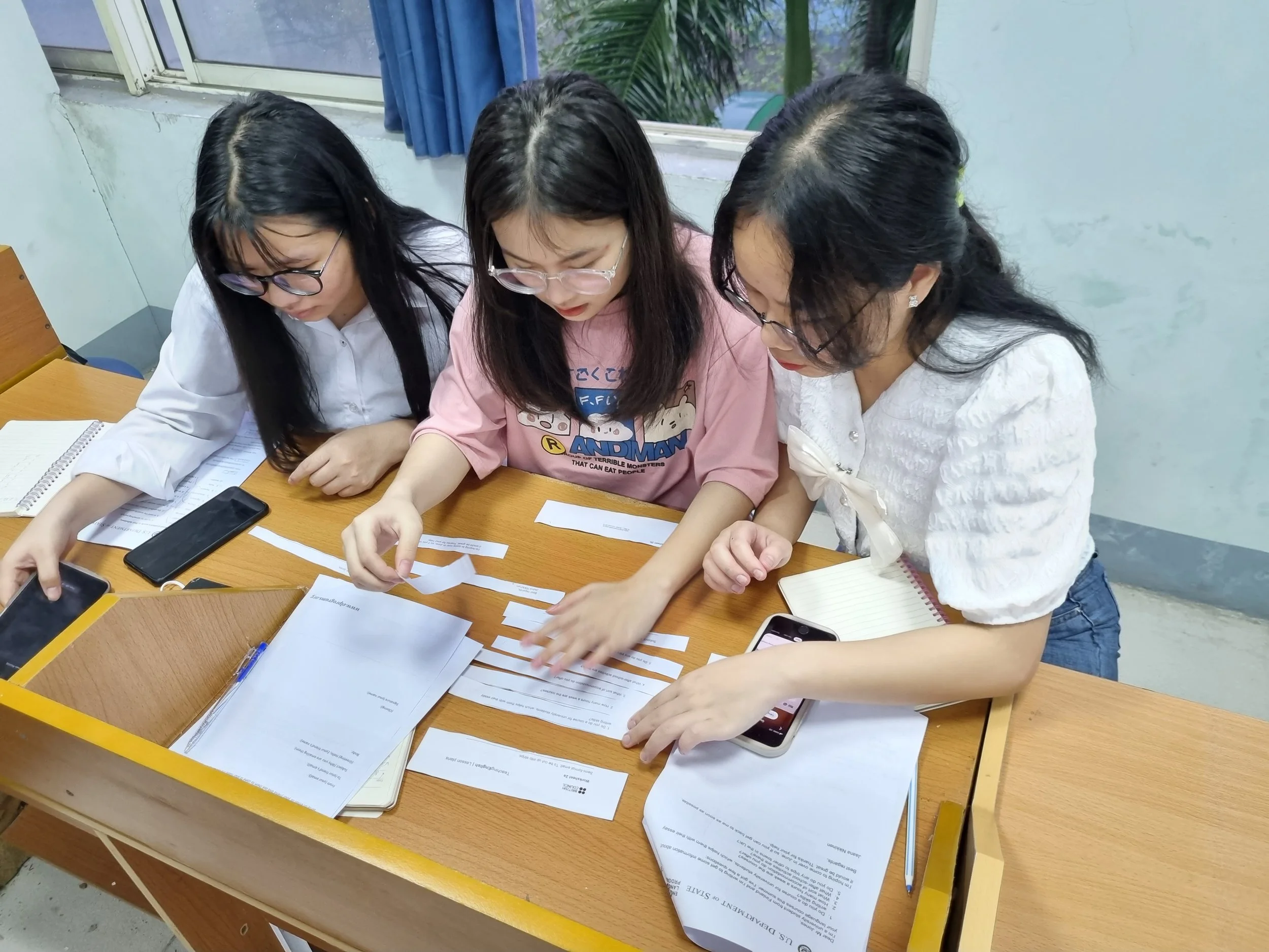 Three women sitting at a table, reviewing papers and working on a project together, with notebooks and smartphones on the table and a window with greenery outside.