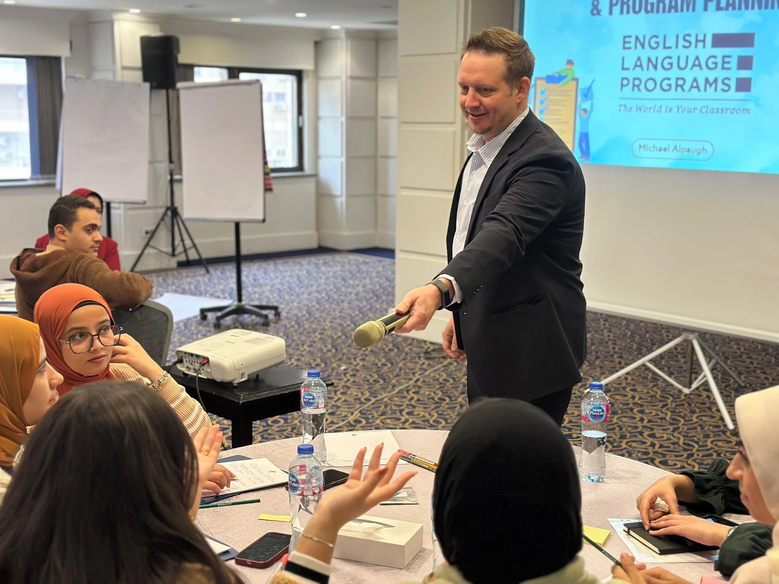 A man in a suit is speaking or presenting to a group of seated individuals in a classroom or conference room. The group includes women wearing headscarves, one woman with glasses, and a man with short hair. There are water bottles and notebooks on the table. A large screen in the background displays a presentation titled 'English Language Programs' by Michael Alpaugh.