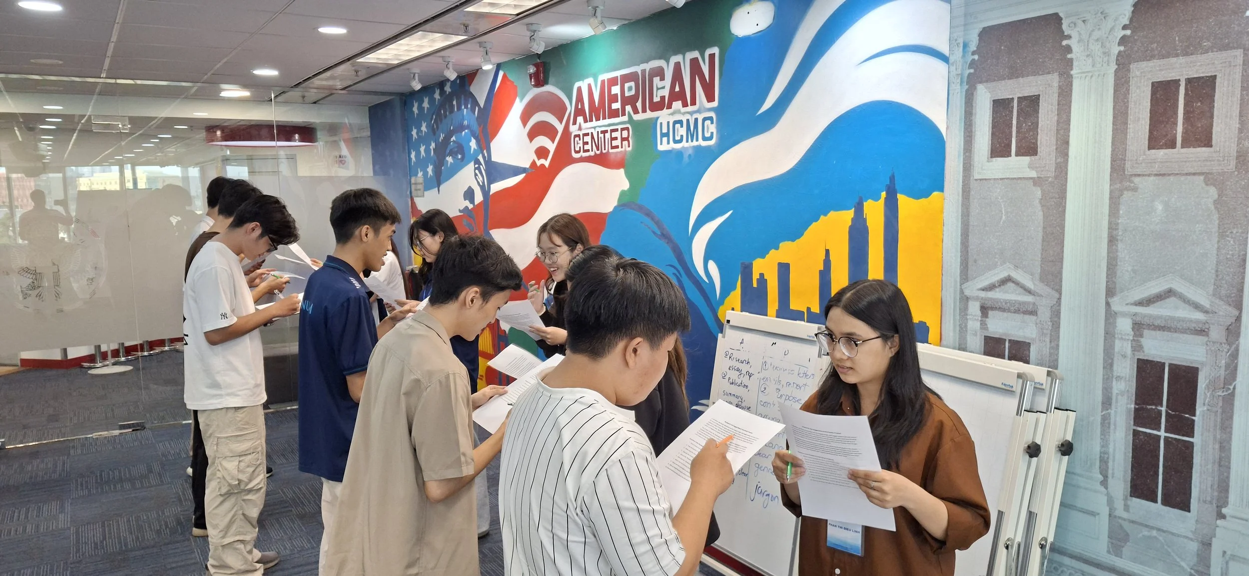 Group of young people gathering around a table with papers in front of a colorful mural at the American Center HCMC.