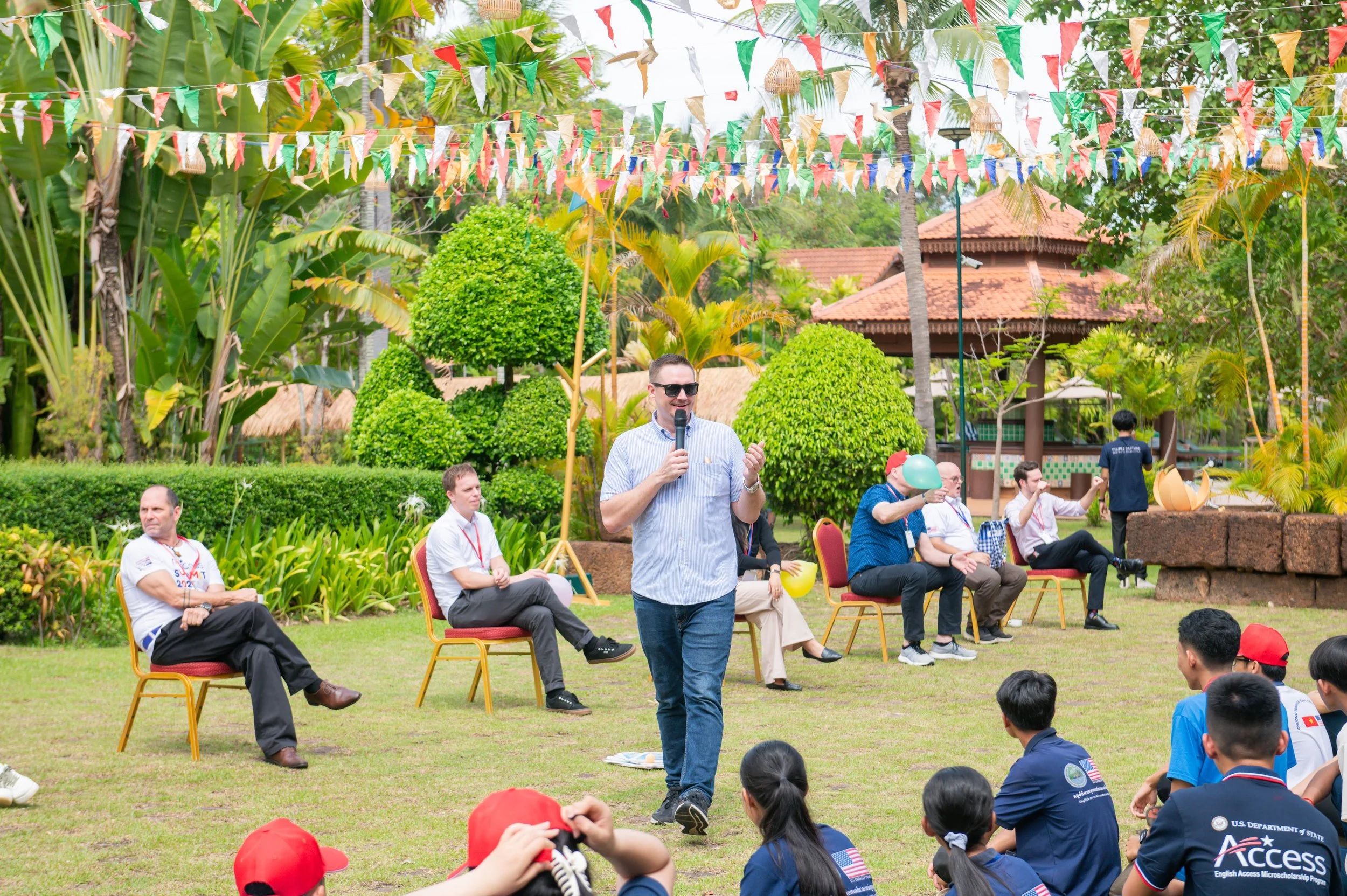 A man in sunglasses and a light blue shirt speaking into a microphone at an outdoor event with people seated on chairs and sitting on the grass. Colorful banners hang above, greenery surrounds the scene, and there are several people wearing dark blue shirts with American flags, red caps, and some balloons.