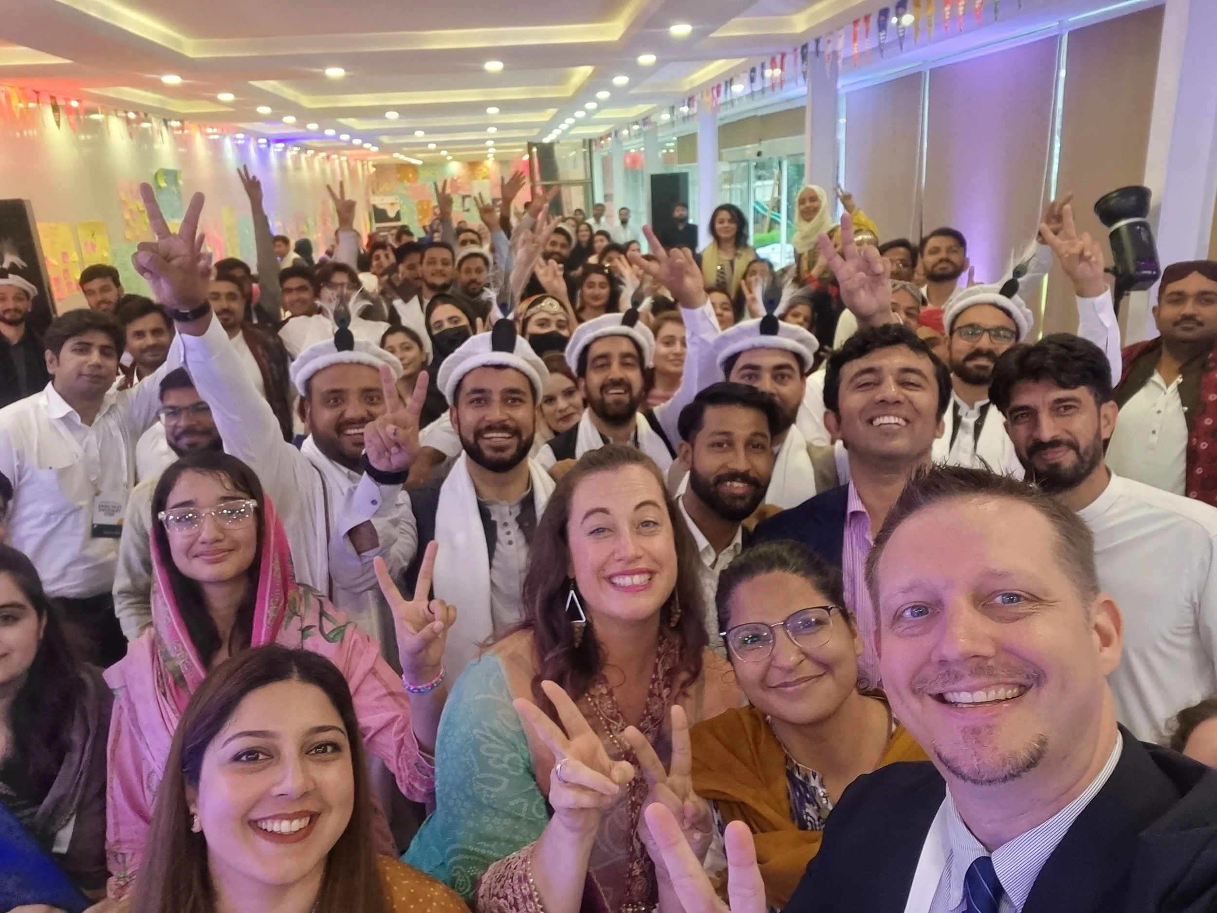 Large group of people at an indoor celebration, many wearing white or colorful traditional clothing, smiling, making peace signs, and looking at the camera. Some people wear headgear, and the background is decorated with colorful banners and lights.
