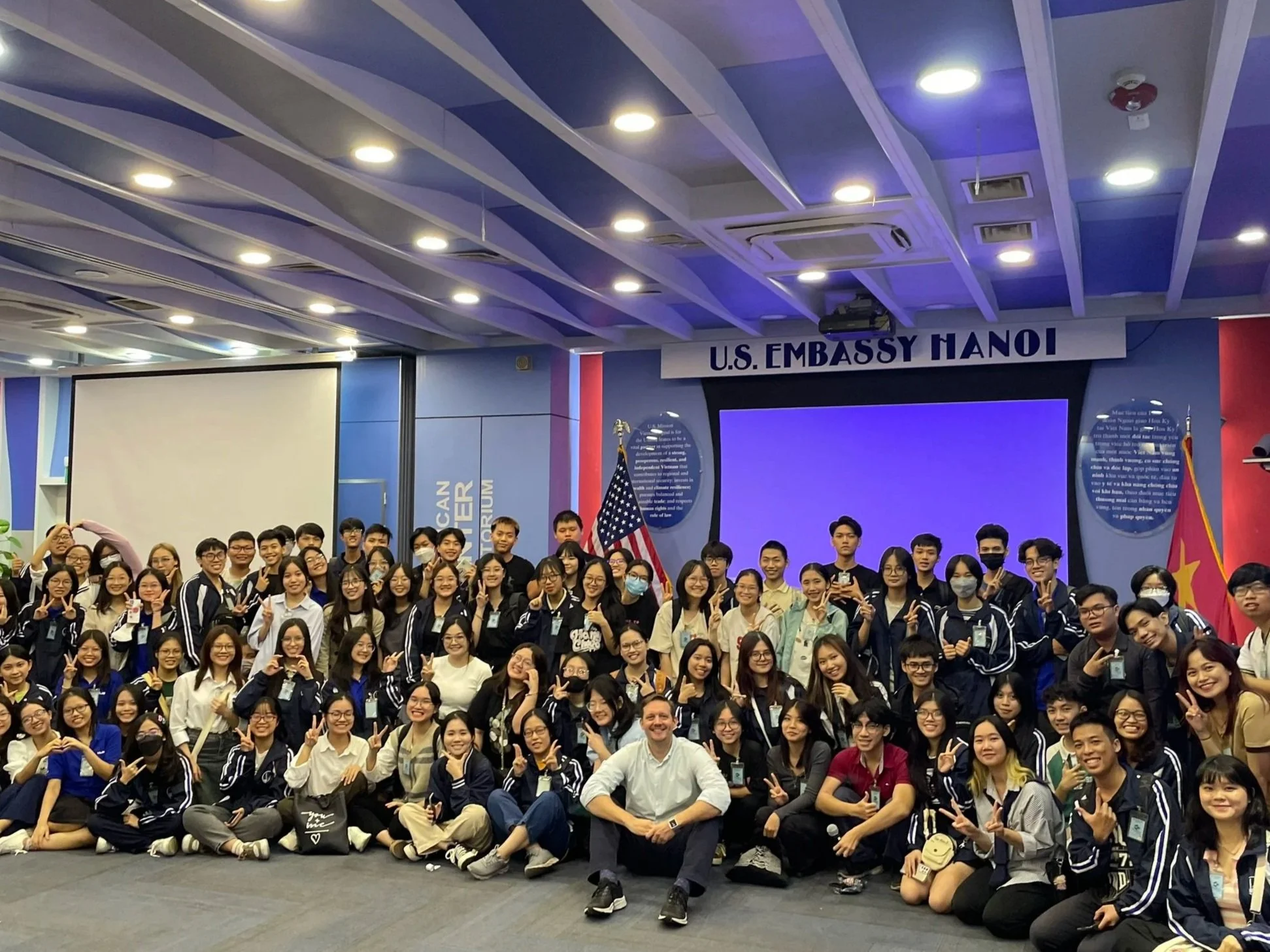 A large group of people posing for a photo in front of a screen at a U.S. Embassy event in Hanoi. The group includes young adults and some older individuals, with many making peace signs and smiling. There are flags and informational banners around, and the setting appears to be an indoor conference or event space with ceiling lights.