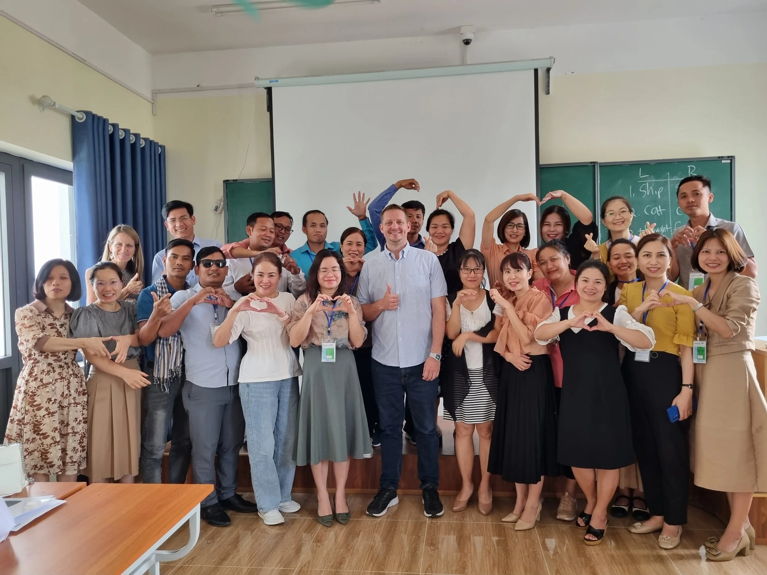 Group of diverse people posing together in a classroom, some making heart shapes with their hands, smiling, with a white projection screen and blackboards in the background.