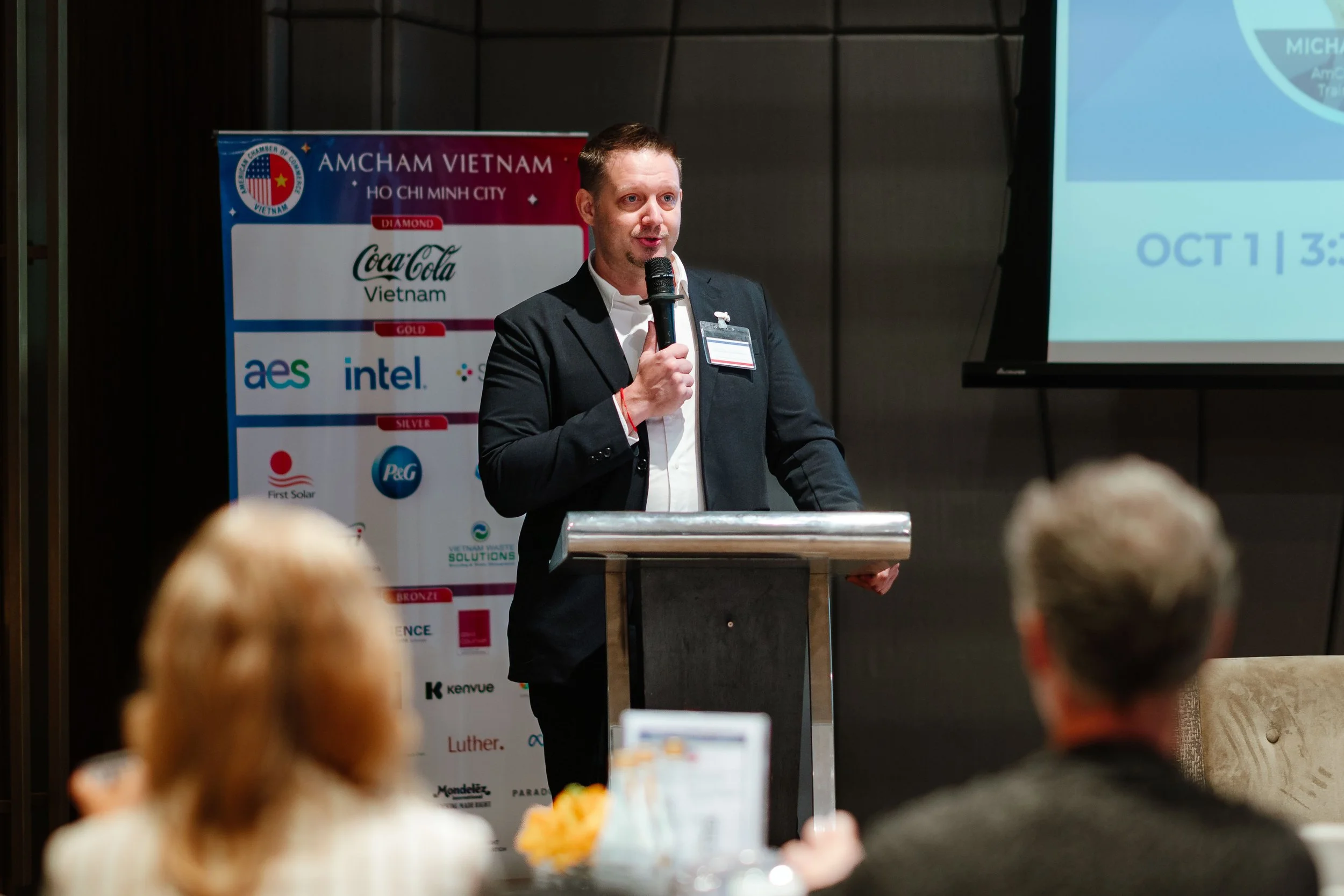 A man in a dark suit speaks into a microphone at a podium during a conference. Behind him is a banner with various logos and text, including 'AmCham Vietnam' and 'Ho Chi Minh City.' A partially visible presentation slide on the right displays the date 'October 13.' Audience members are seated and listening.