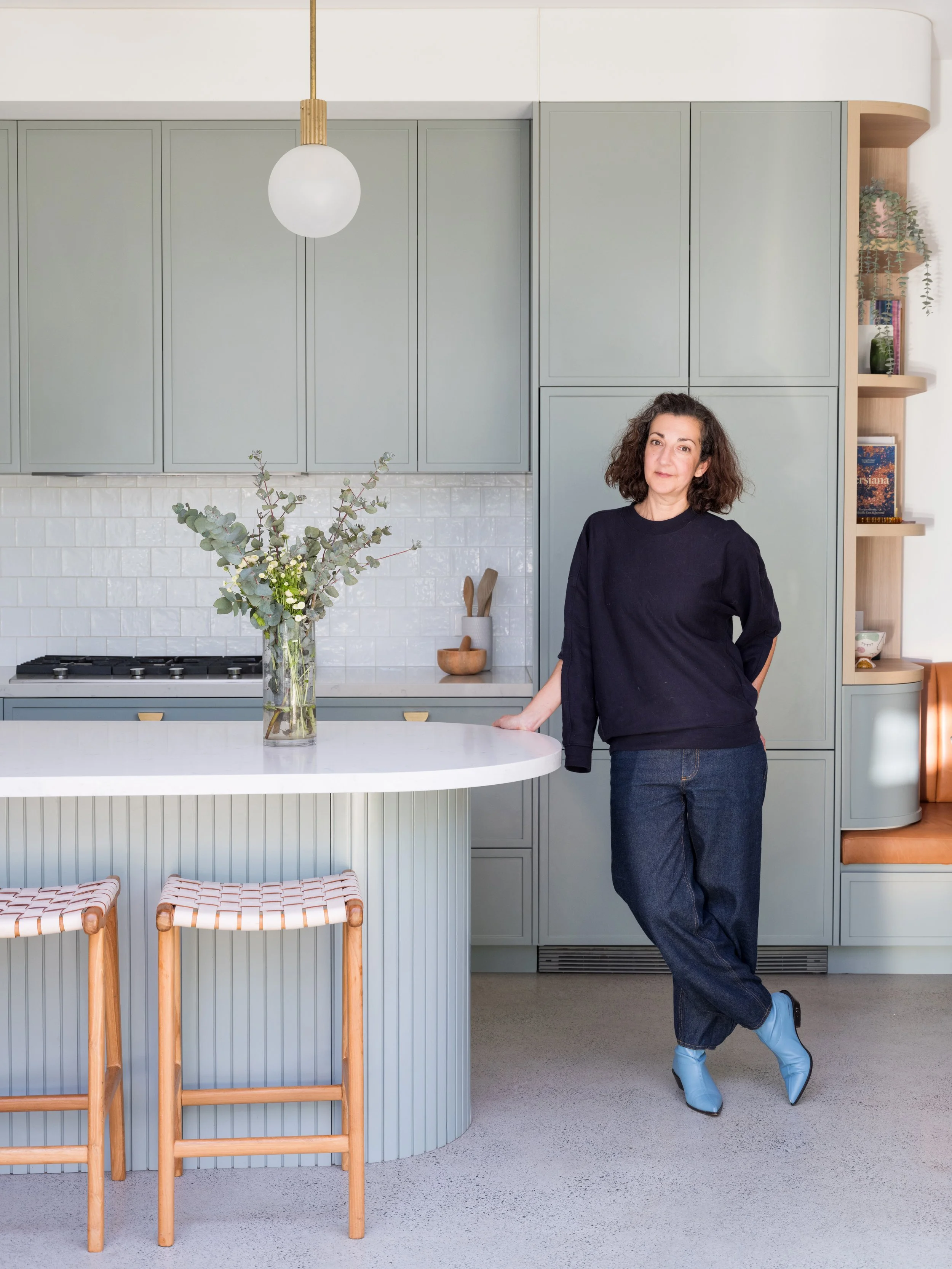 Woman standing in a modern kitchen with green cabinets, a white countertop, and a vase with eucalyptus and white flowers. She has curly brown hair, is wearing a black top, jeans, blue boots, and is leaning against the counter.