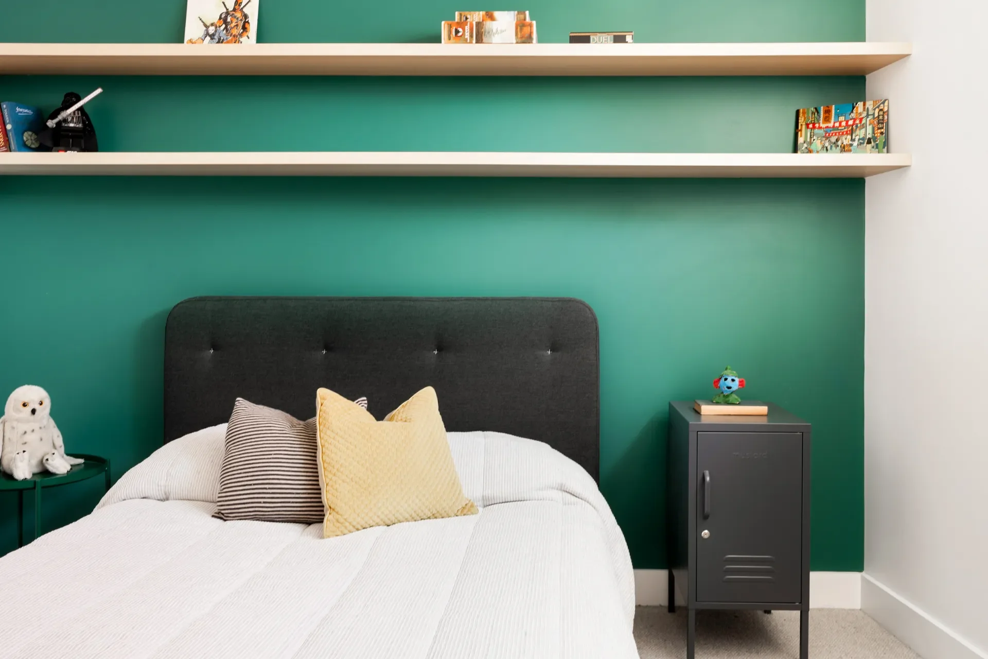 A bedroom with a green accent wall, a bed with a white quilt and decorative pillows, a gray nightstand with a small blue robot toy on top, and two white floating shelves holding books and artwork.