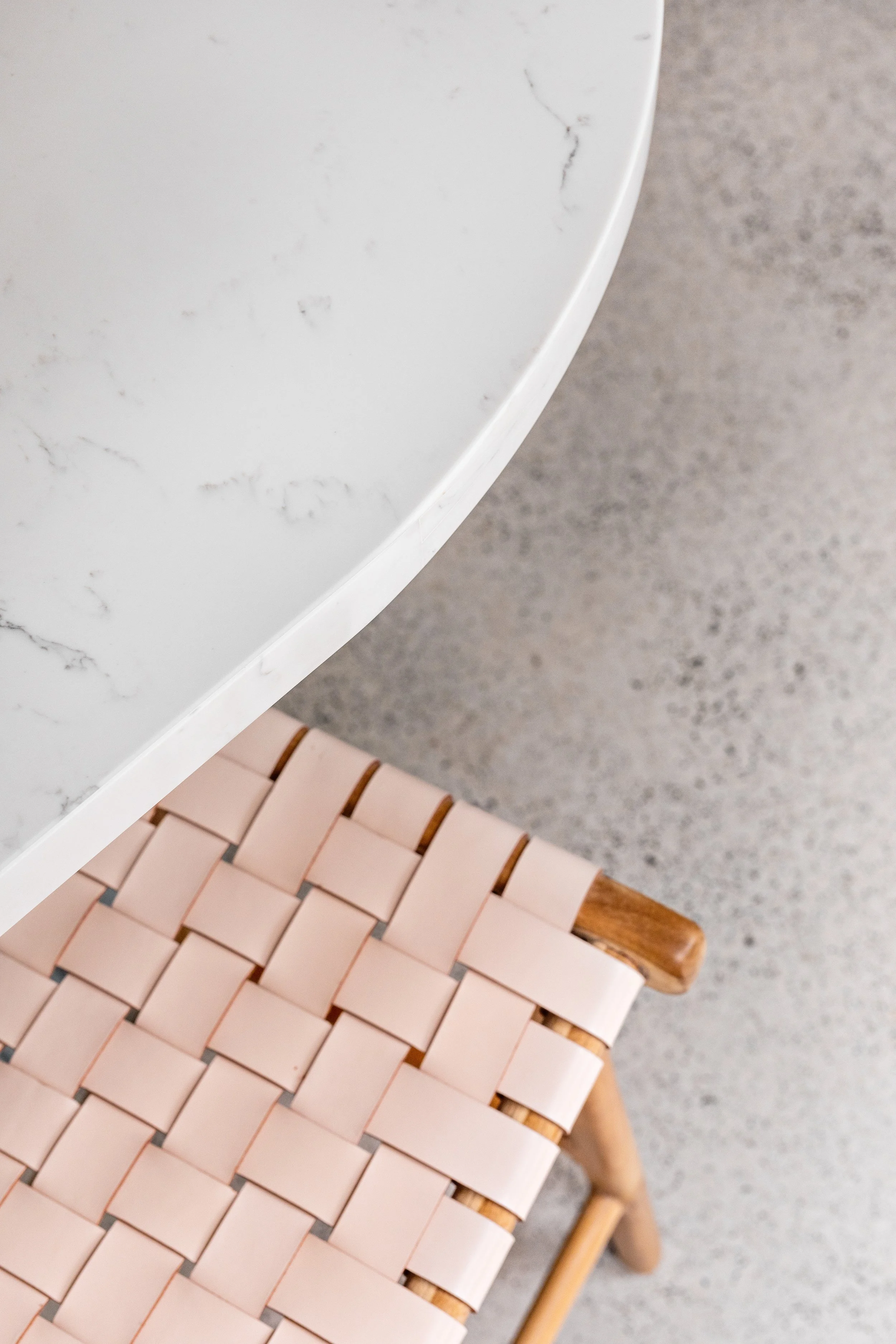 Close-up of a white marble table with visible veins against a gray concrete floor, and part of a chair with pink woven surface and a wooden armrest.