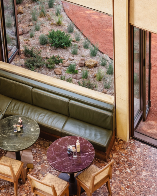 Indoor cafe with a green leather booth, two marble-topped tables, and four wooden chairs. Large windows and a door open to an outdoor patio with desert landscaping and rocks.