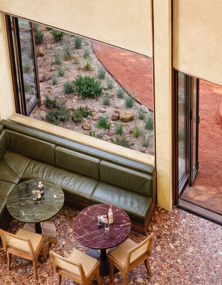 A cozy indoor seating area with a green leather banquette, two round marble-top tables, and four wooden chairs. Outside, a garden with rocks and desert plants is visible through large windows and an open sliding glass door.