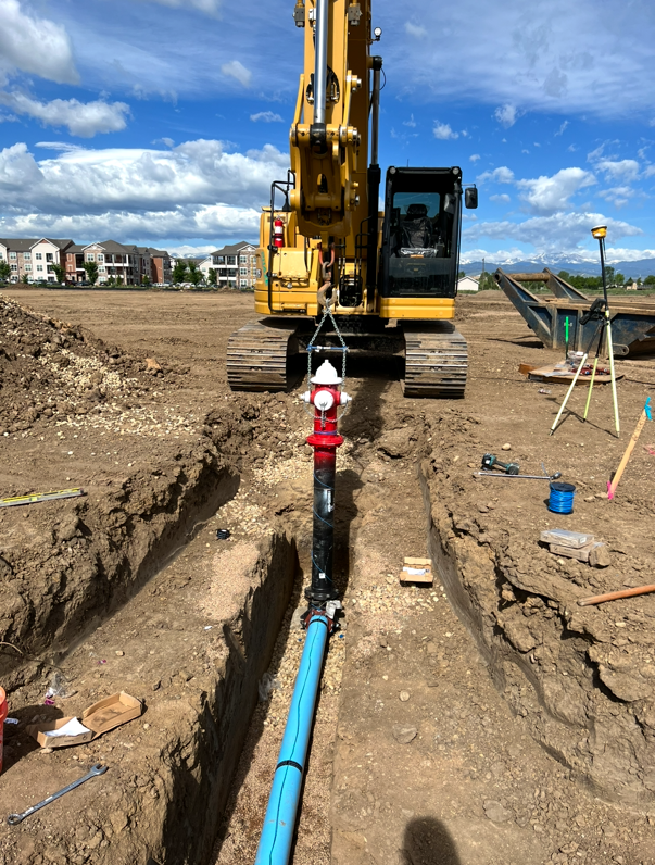 Construction site with a yellow excavator installing a water pipe and fire hydrant in a trench, with modern apartment buildings in the background and clear blue skies.