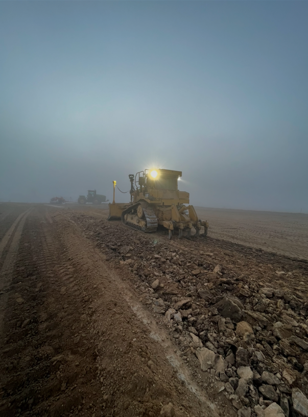 A bulldozer grading a dirt road at sunrise or sunset, with another machinery in the background.