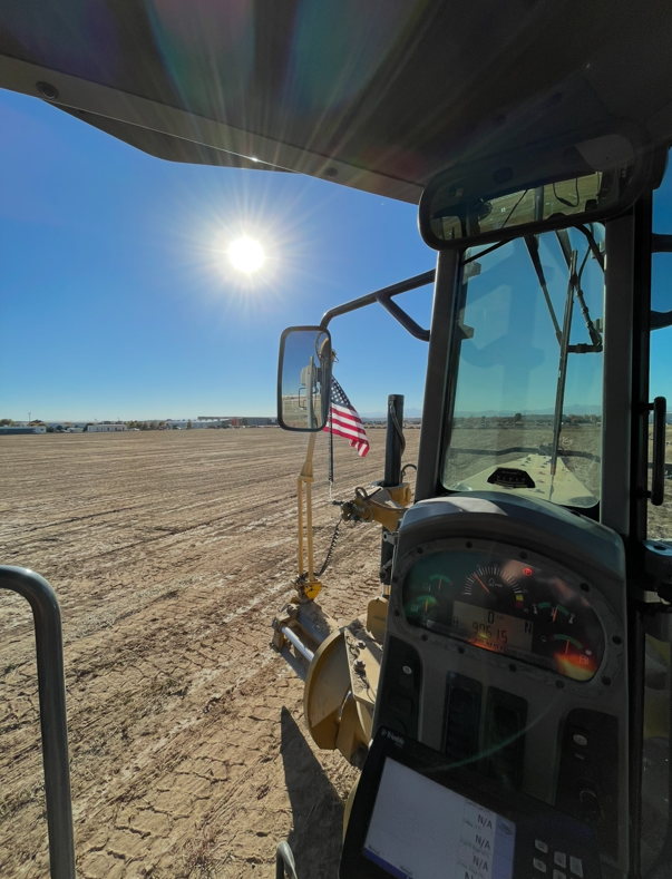 View from inside a construction or farming vehicle cab showing a sunlit dirt field, an American flag, and a clear sky with the sun shining brightly.
