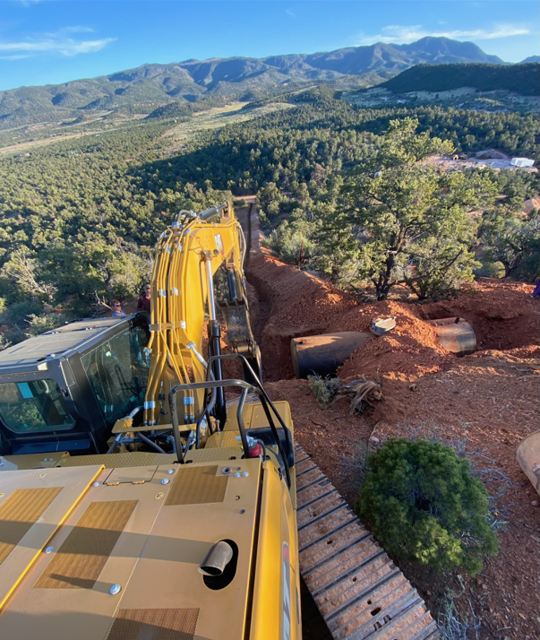 A yellow excavator working on a construction or excavation site amid a natural landscape with trees and mountain views.