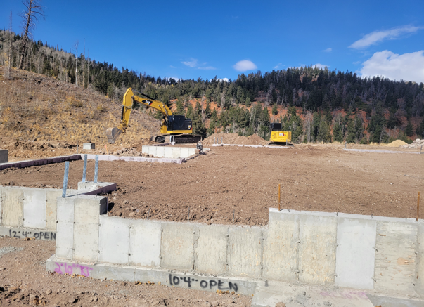 Construction site with an excavator and a generator on a dirt field, surrounded by a concrete wall, with hills and trees in the background under a blue sky.