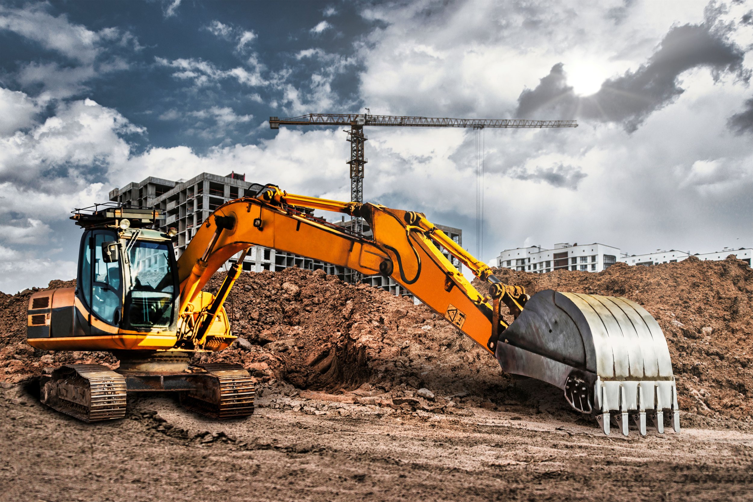A yellow excavator digging into dirt at a construction site, with an overcast sky and a crane and buildings in the background.