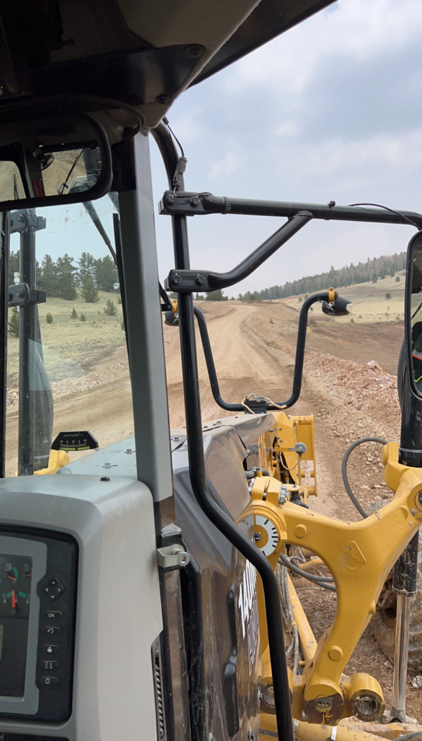 View from inside a construction vehicle cab showing a dirt road ahead and rolling hills in the background.