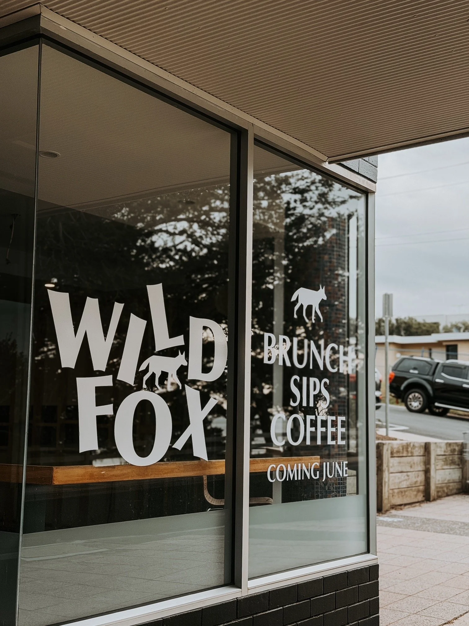 A storefront window with promotional text including 'WILD', 'FOX', 'BRUNCH', 'SIPS', 'COFFEE', and 'COMING JUNE' with a silhouette of a fox and a wolf.
