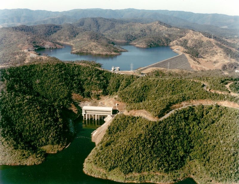 A landscape view of a dam and reservoir surrounded by hills with green vegetation in a rural area.