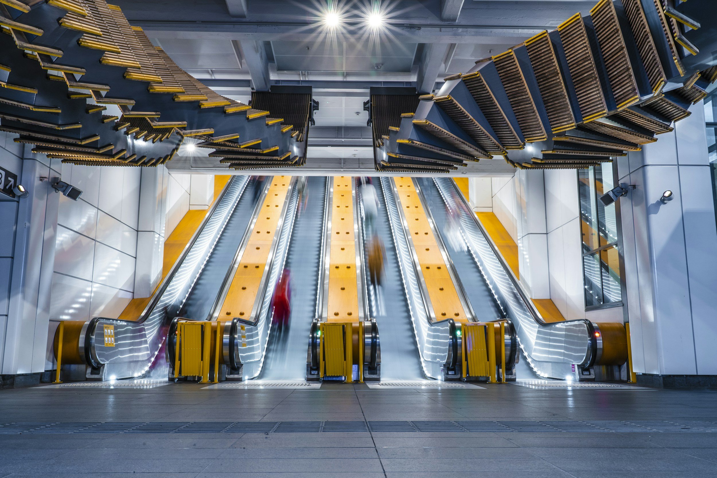 Underground escalator station with two moving escalators, yellow accents, modern design, bright overhead lighting, and a person blurred in red walking down one escalator.