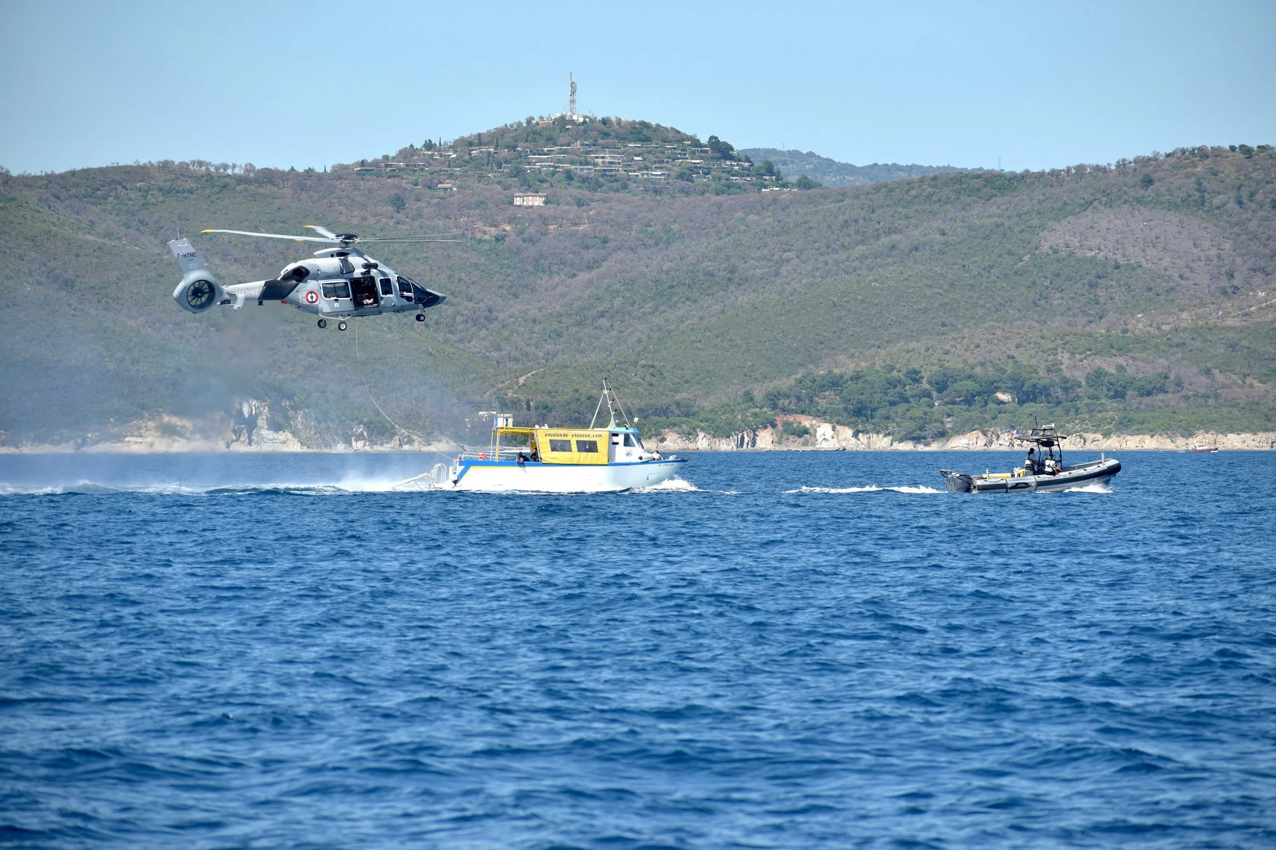 A helicopter flying over water with two boats below, a mountainous landscape with buildings and a communication tower in the background.