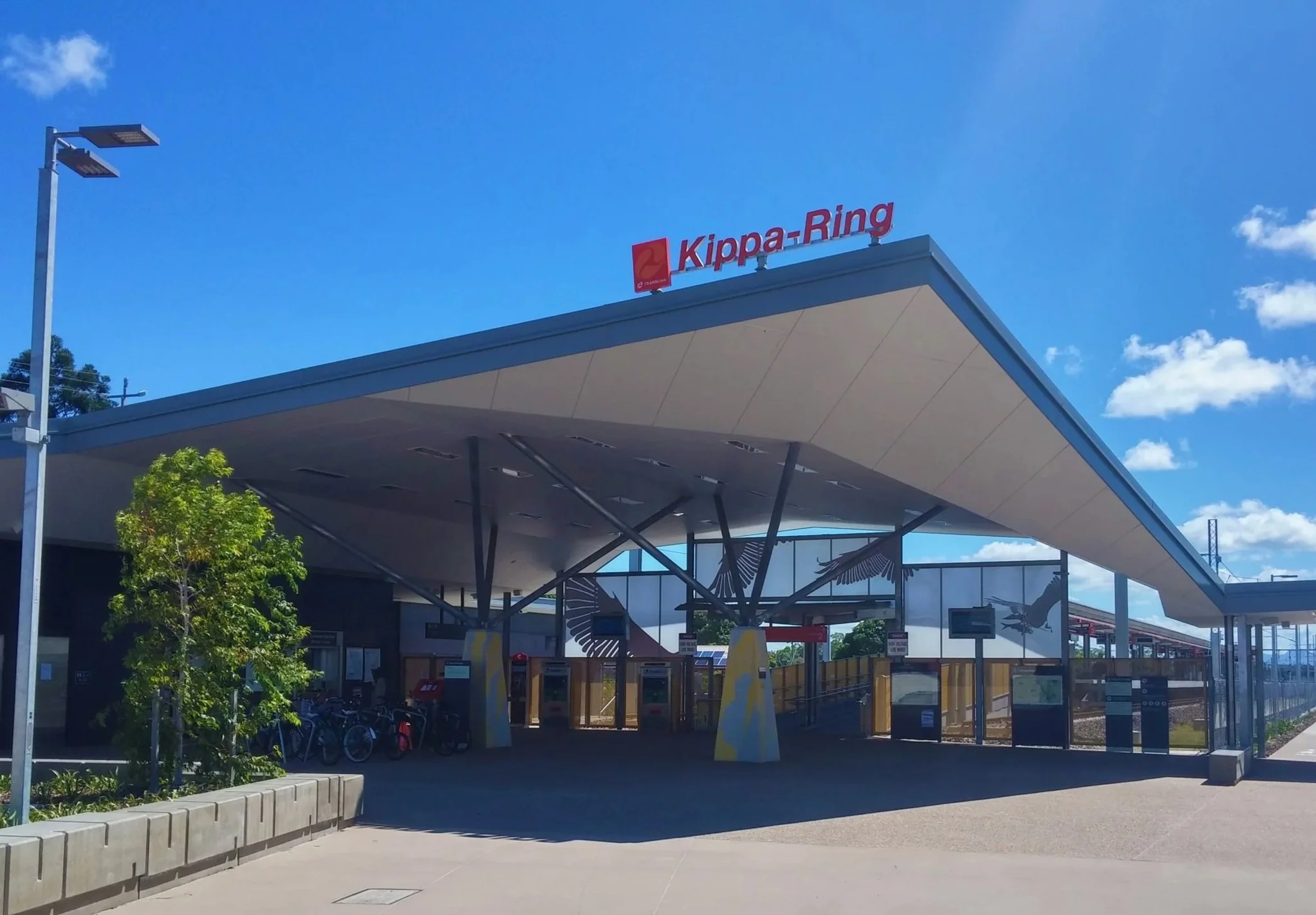 The image shows a modern train station with a sign that reads 'Kippa-Ring' on a building. The station has a distinctive large, curved roof supported by sleek metal beams. There are trees and bicycles visible, and the sky is clear with some clouds.