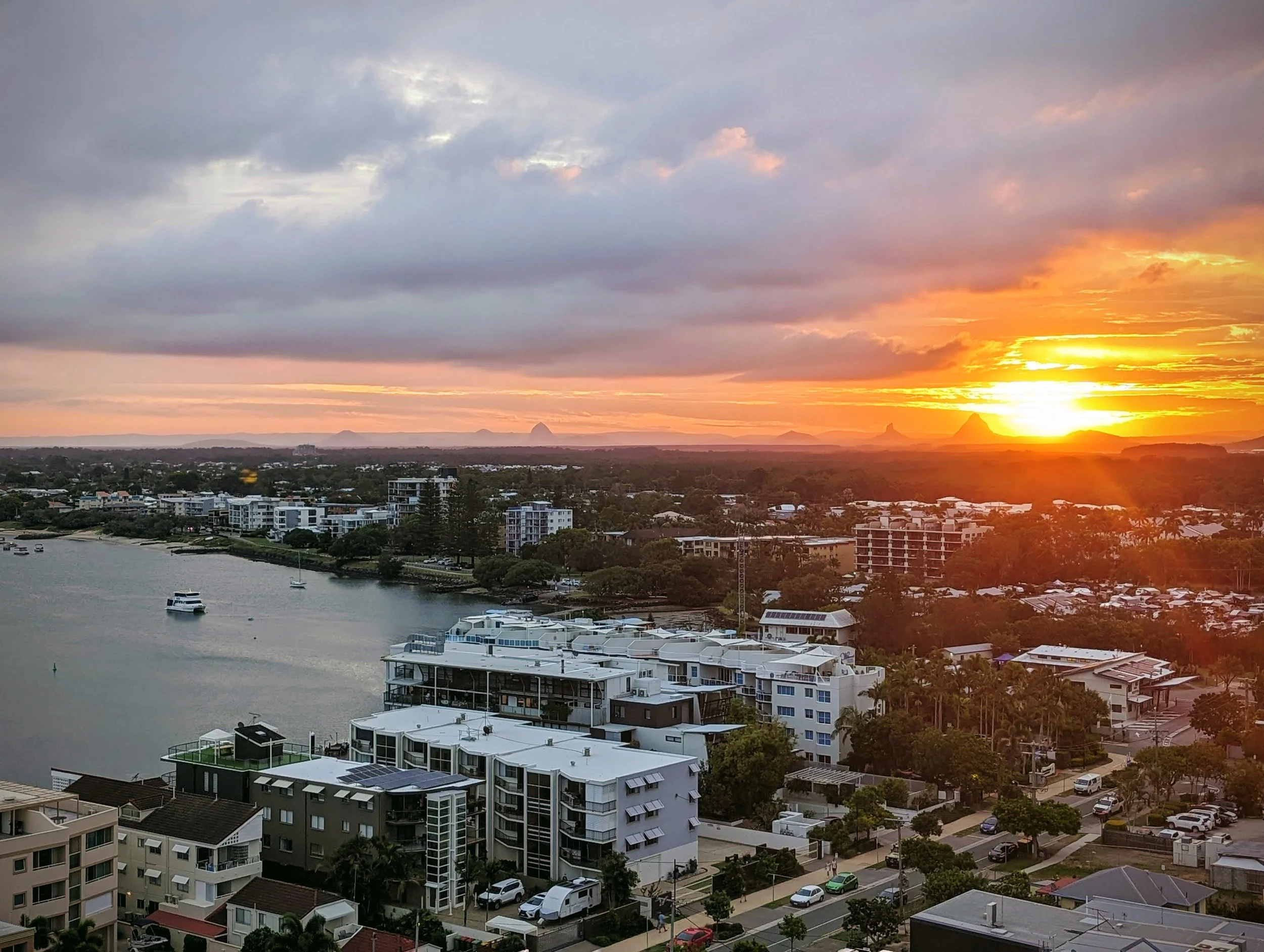 A city waterfront at sunset with buildings, boats in the water, and a sky filled with clouds and a bright orange sun.