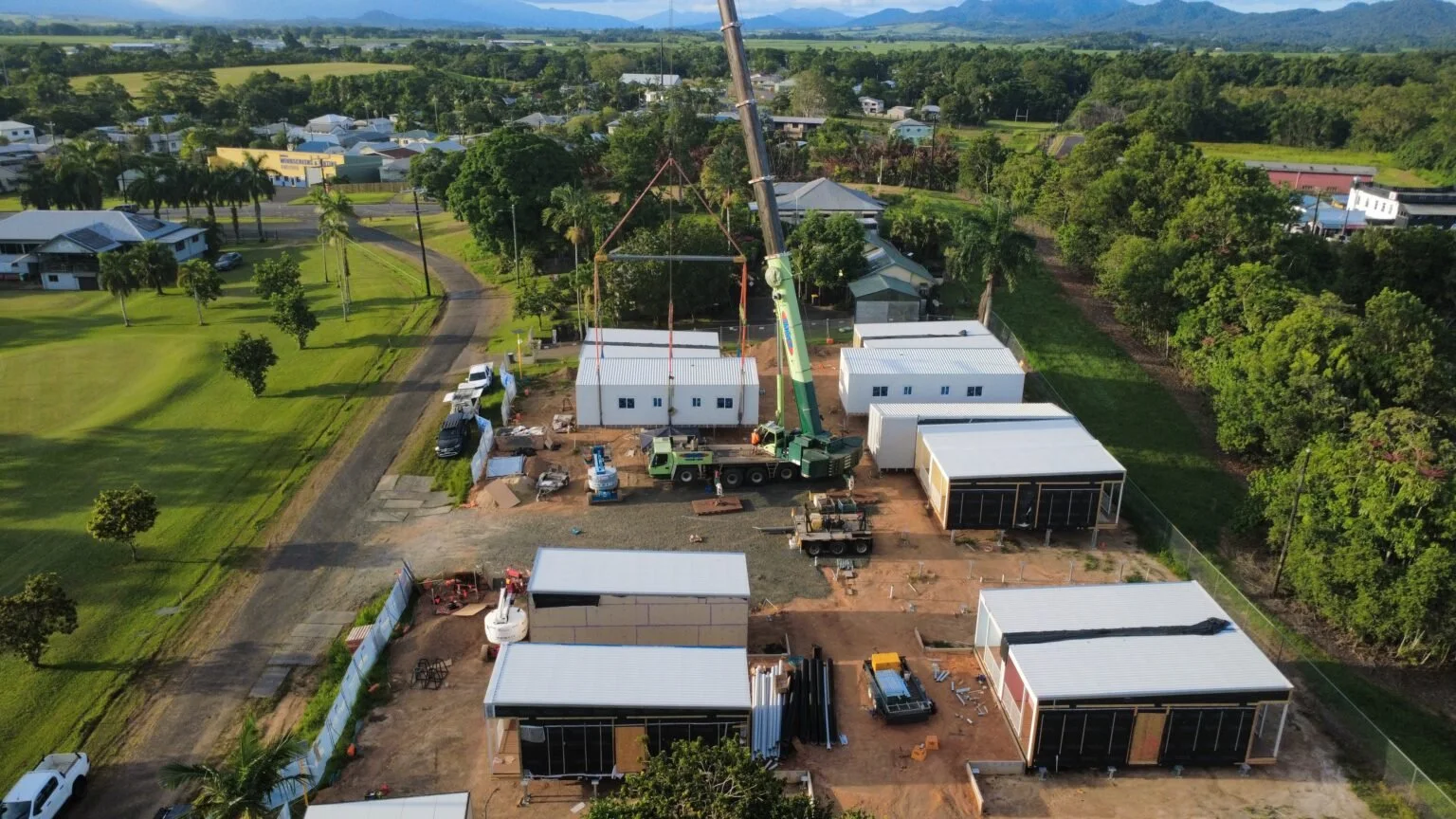 Aerial view of a construction site with a crane lifting a structure, multiple modular buildings, construction vehicles, and green surrounding landscape.