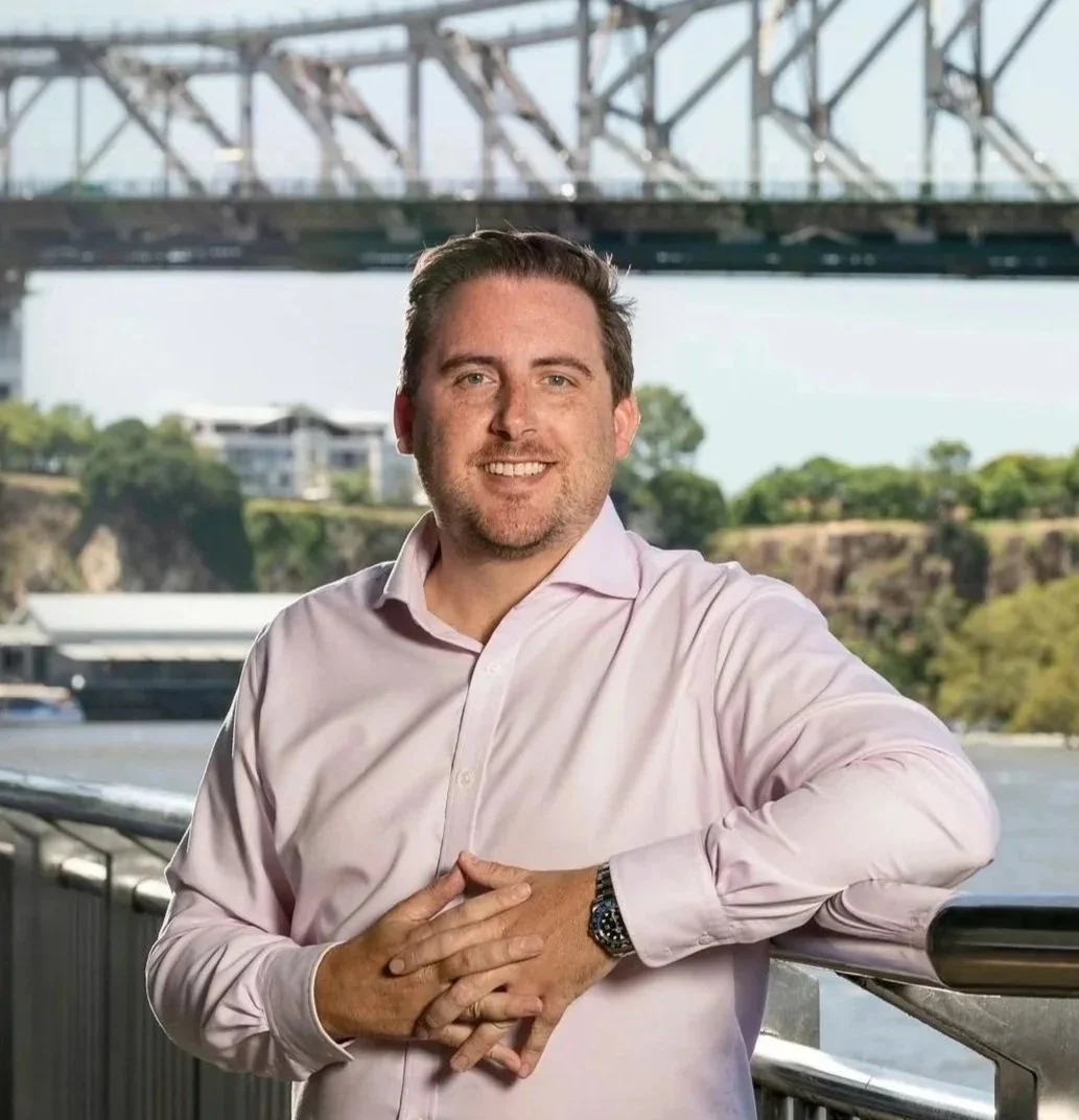 A man smiling with a bridge, water, and green trees in the background, standing on a railing outdoors.