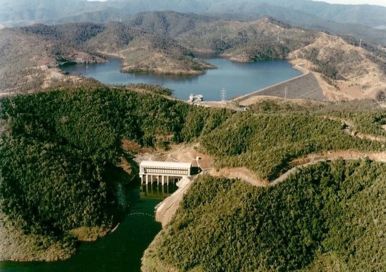 A large dam with a concrete spillway surrounded by green hills and a reservoir behind it.