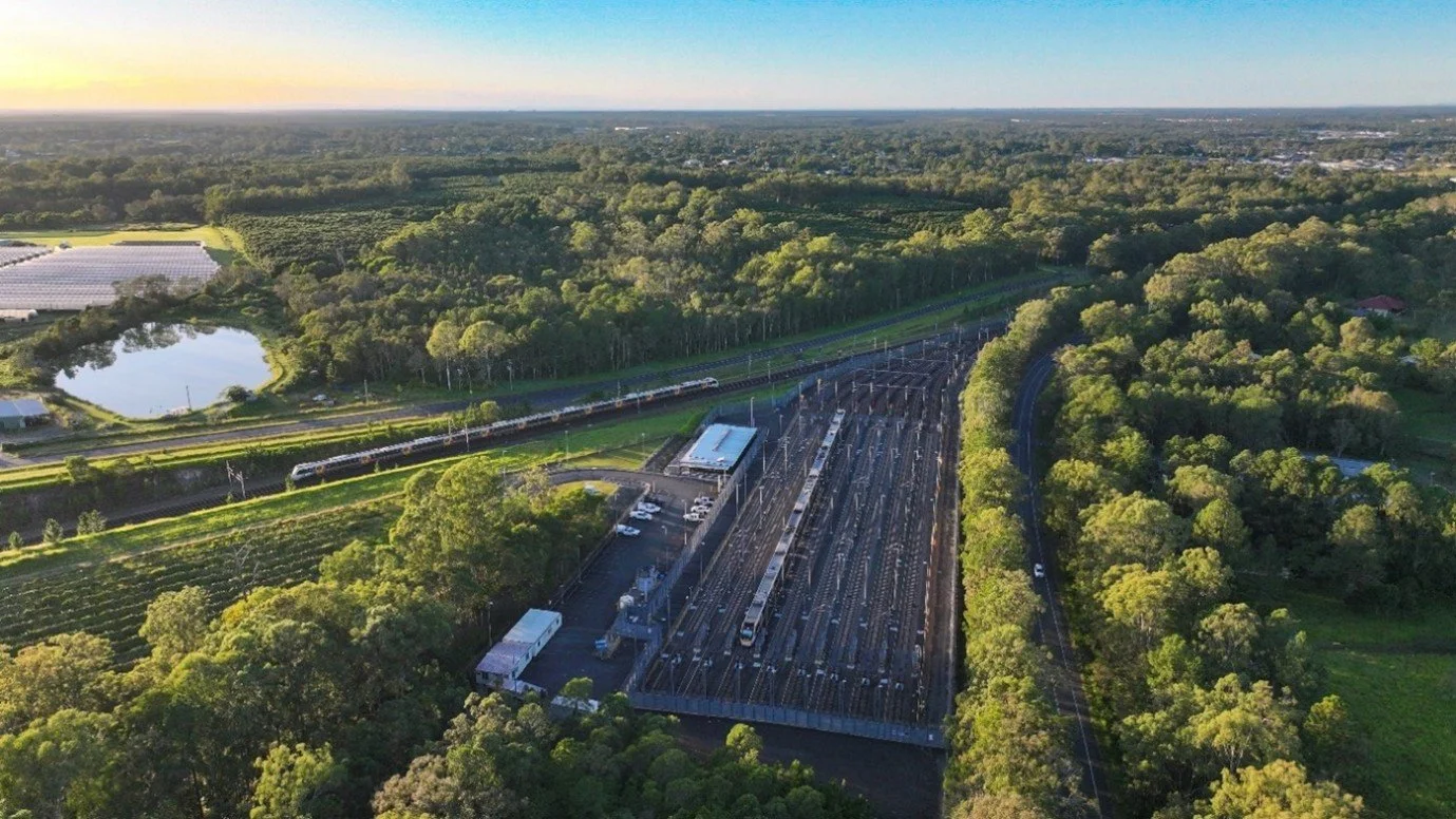 Aerial view of a train station with multiple train tracks, trains parked, surrounded by greenery and trees, with a pond and fields in the background under a clear sky.