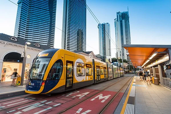 Modern yellow tram on city street with tall skyscrapers in the background and passengers waiting at the station.