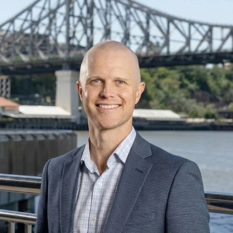 Man in a gray blazer and checked shirt smiling outdoors near a river with a bridge in the background.