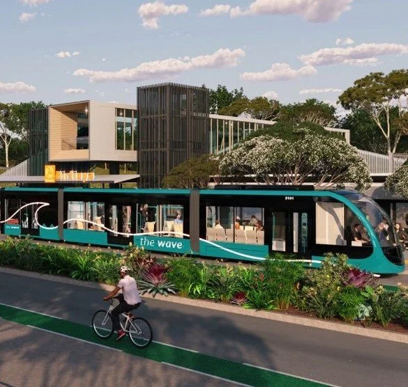 A modern light blue and black tram with the words 'the wave' on its side, traveling on a dedicated bike lane with a cyclist riding beside it. In the background, there is a contemporary building with large windows and wooden accents, surrounded by trees and greenery.