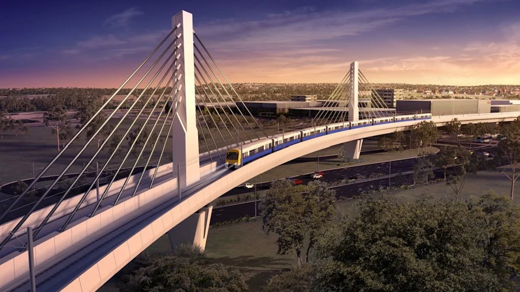 A modern cable-stayed bridge with a train traveling across, surrounded by trees and a parking lot, during sunset.