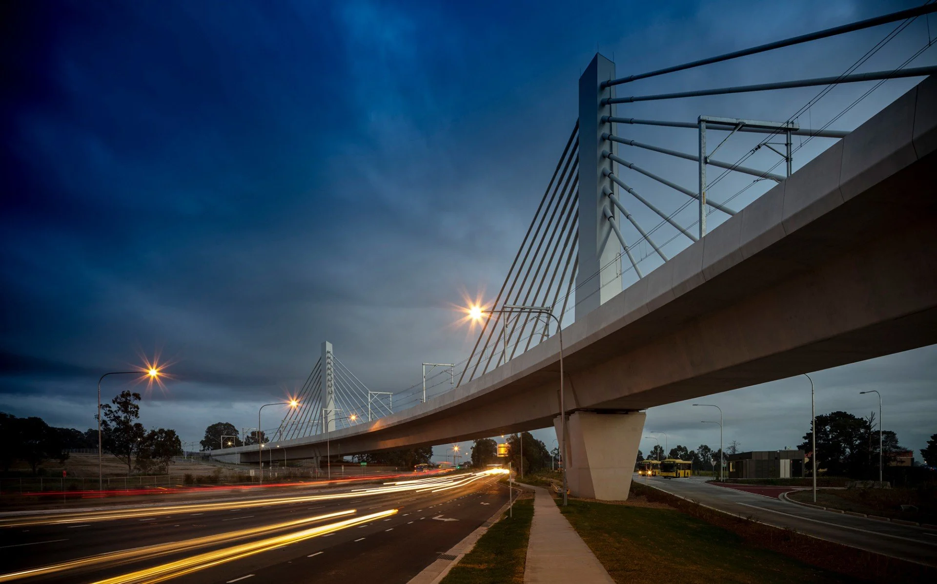A modern cable-stayed bridge during twilight with street lights and light trails from moving vehicles below, trees and a bus in the background.