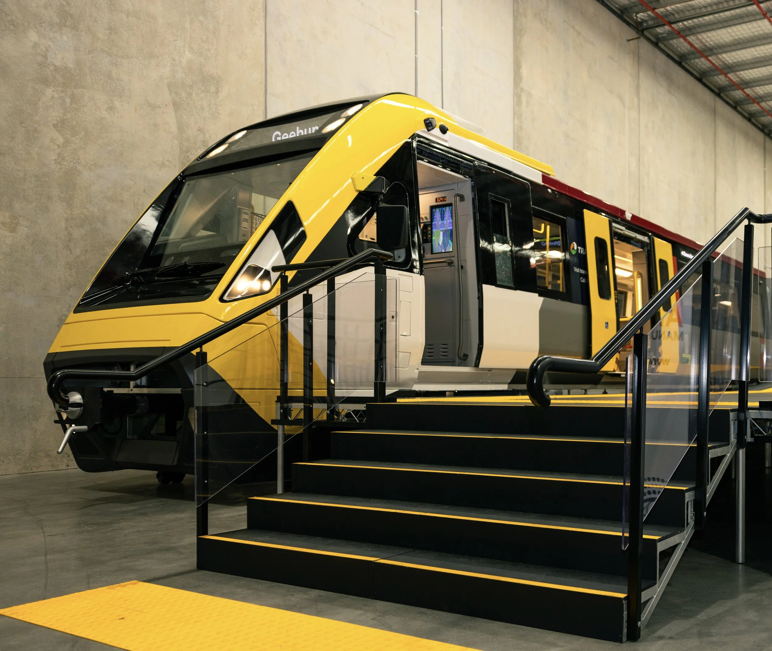 A modern yellow and black tramcar inside a station, with gray walls and a yellow tactile paving at the base of the stairs.