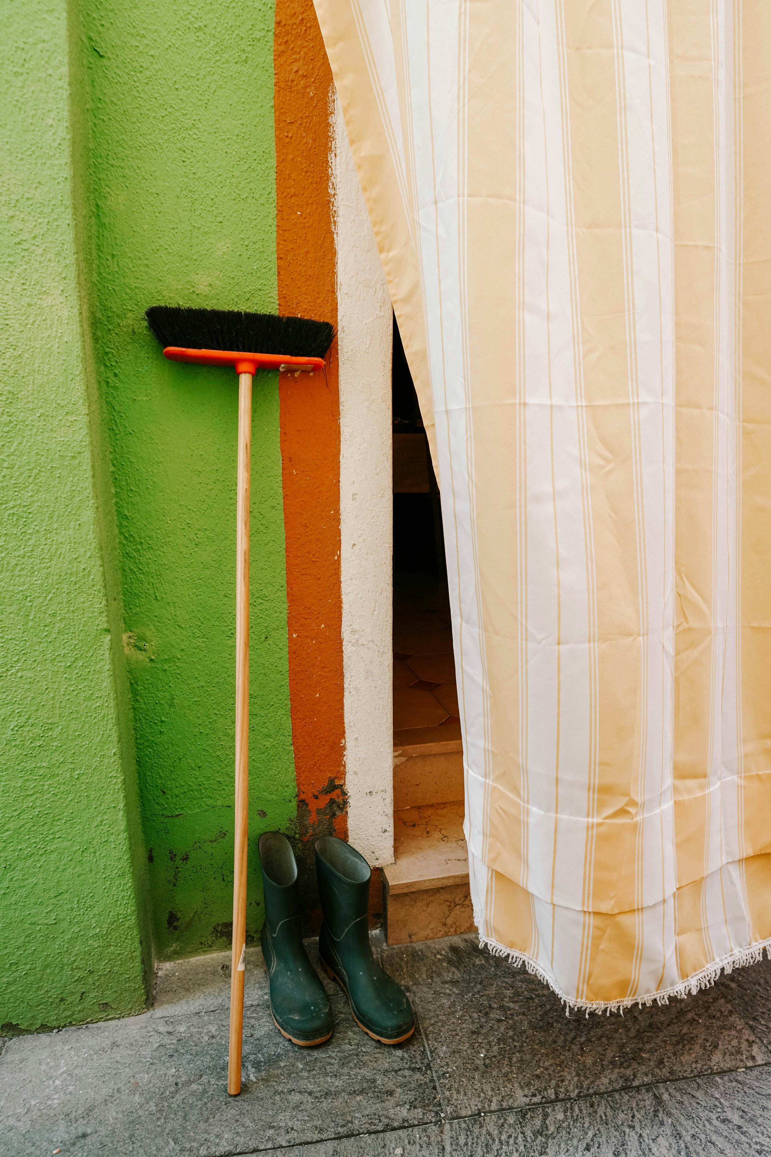 A broom leaning against a green wall, a pair of green rain boots, and a pair of black shoes on the ground. A striped curtain partially covers an entrance.