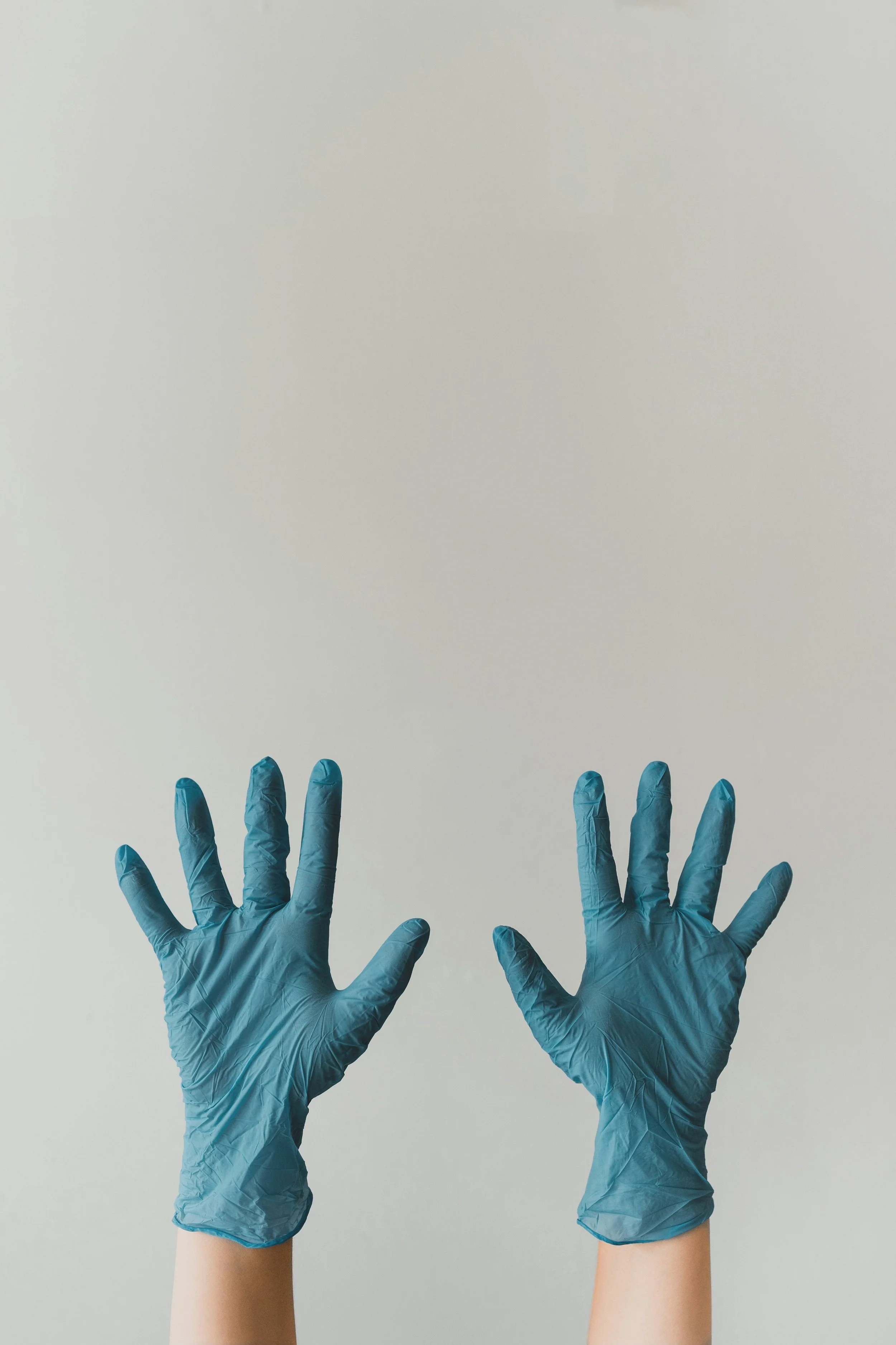 Two hands wearing blue disposable gloves are reaching up against a plain, light-colored background.