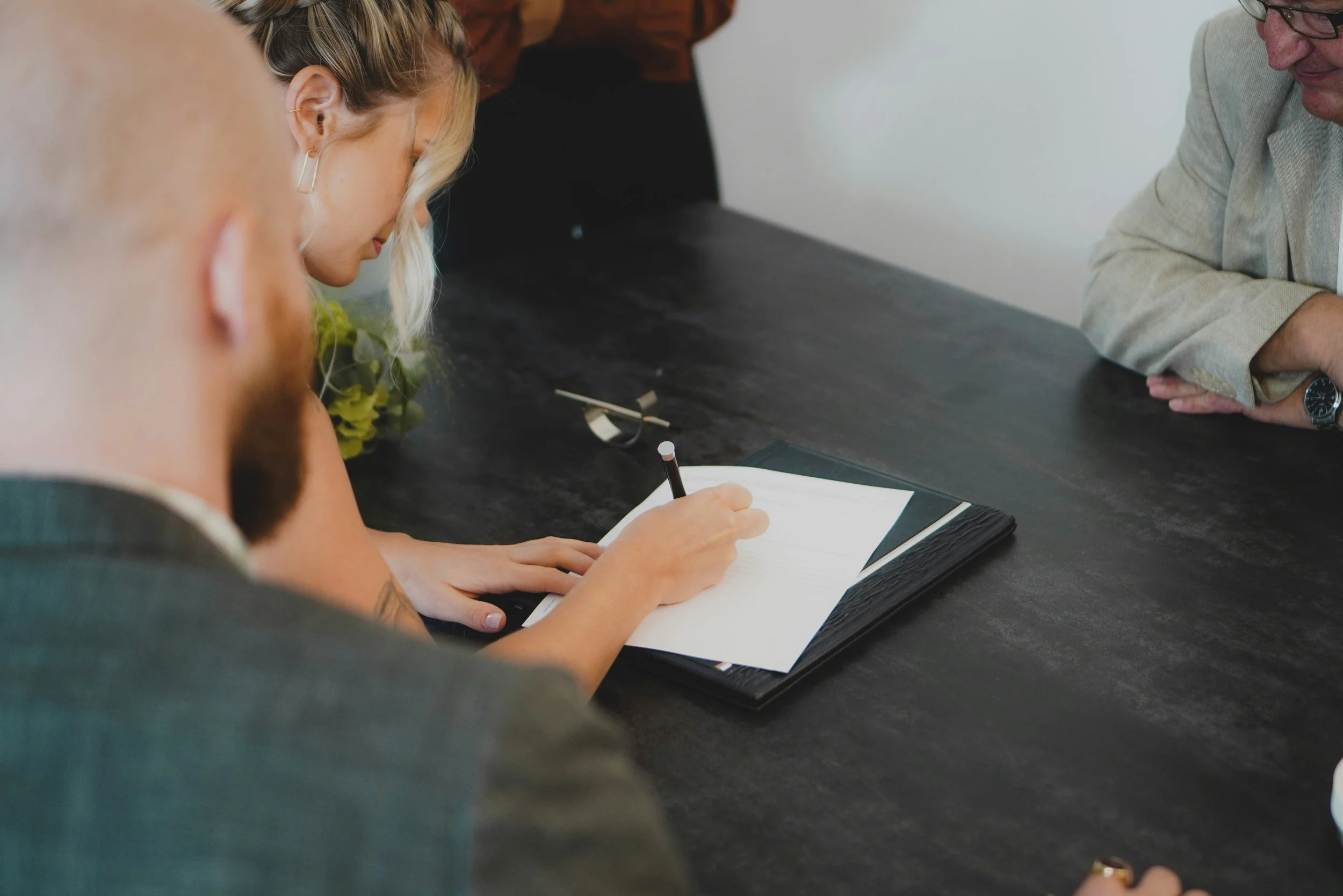 A group of people sitting around a table, engaged in a signing or agreement process. One person is writing in a notebook, while others observe.