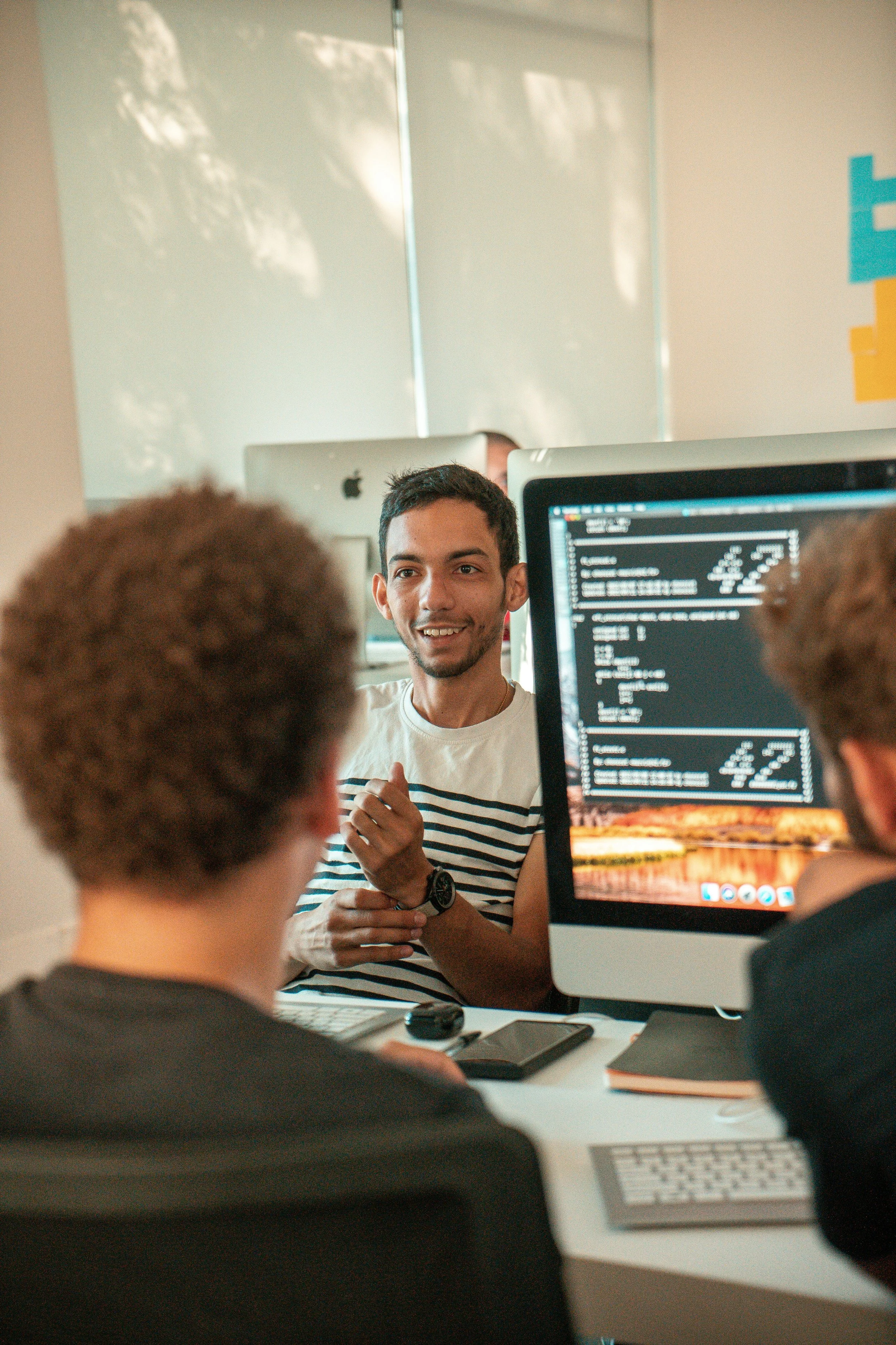 A group of people in a meeting room, with one person speaking and others listening, using desktop computers and laptops.