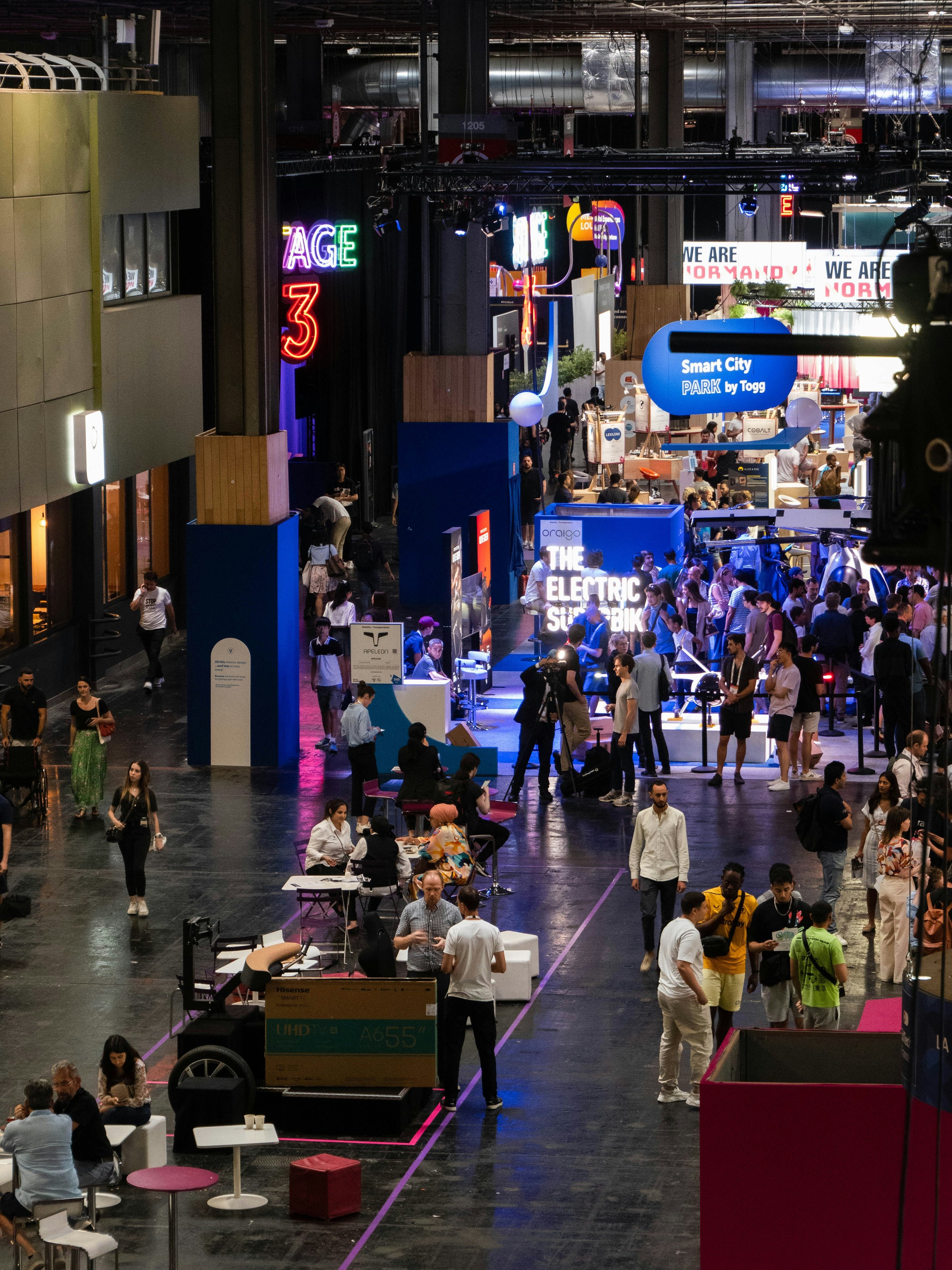 A busy indoor exhibition or convention space with various booths and displays. People are walking, talking, and sitting at tables. Colorful neon signs and digital screens are visible, including signs that read 'Smart City' and 'The Electric Submix.' There are modern furniture and technology demonstrations, with some attendees engaging with exhibitors.