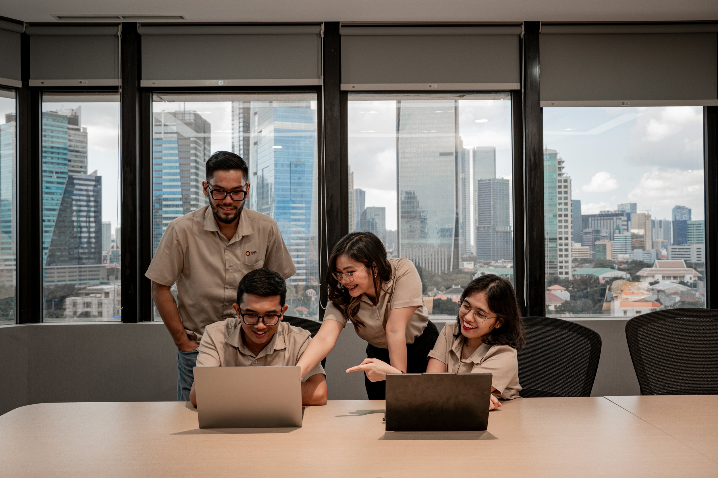 Group of five young professionals, three women and two men, gathered around a table with laptops in a modern office with large windows showing a city skyline.