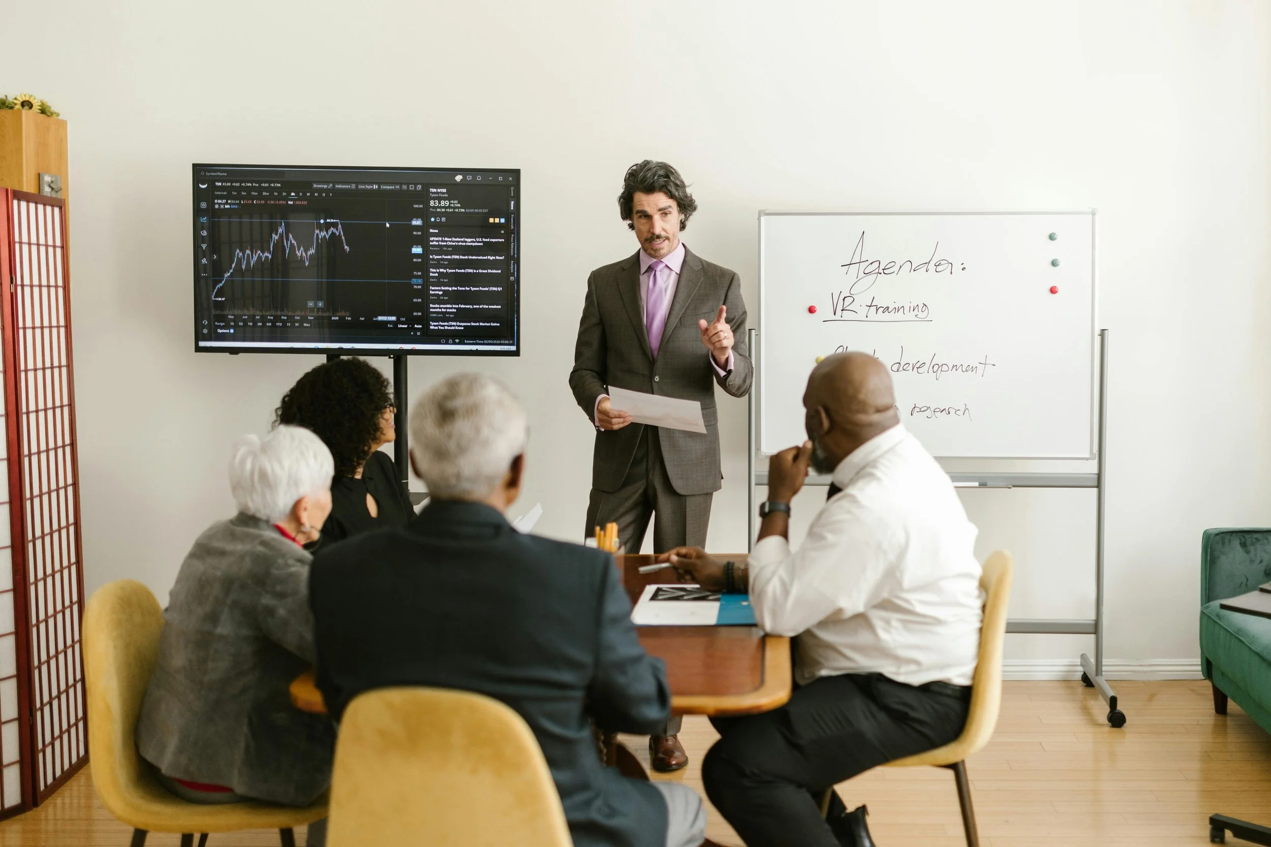 A man in a gray suit giving a presentation in a meeting room with a TV displaying stock market data, and a whiteboard with agenda items including VR training. Several attendees are seated facing the presenter.