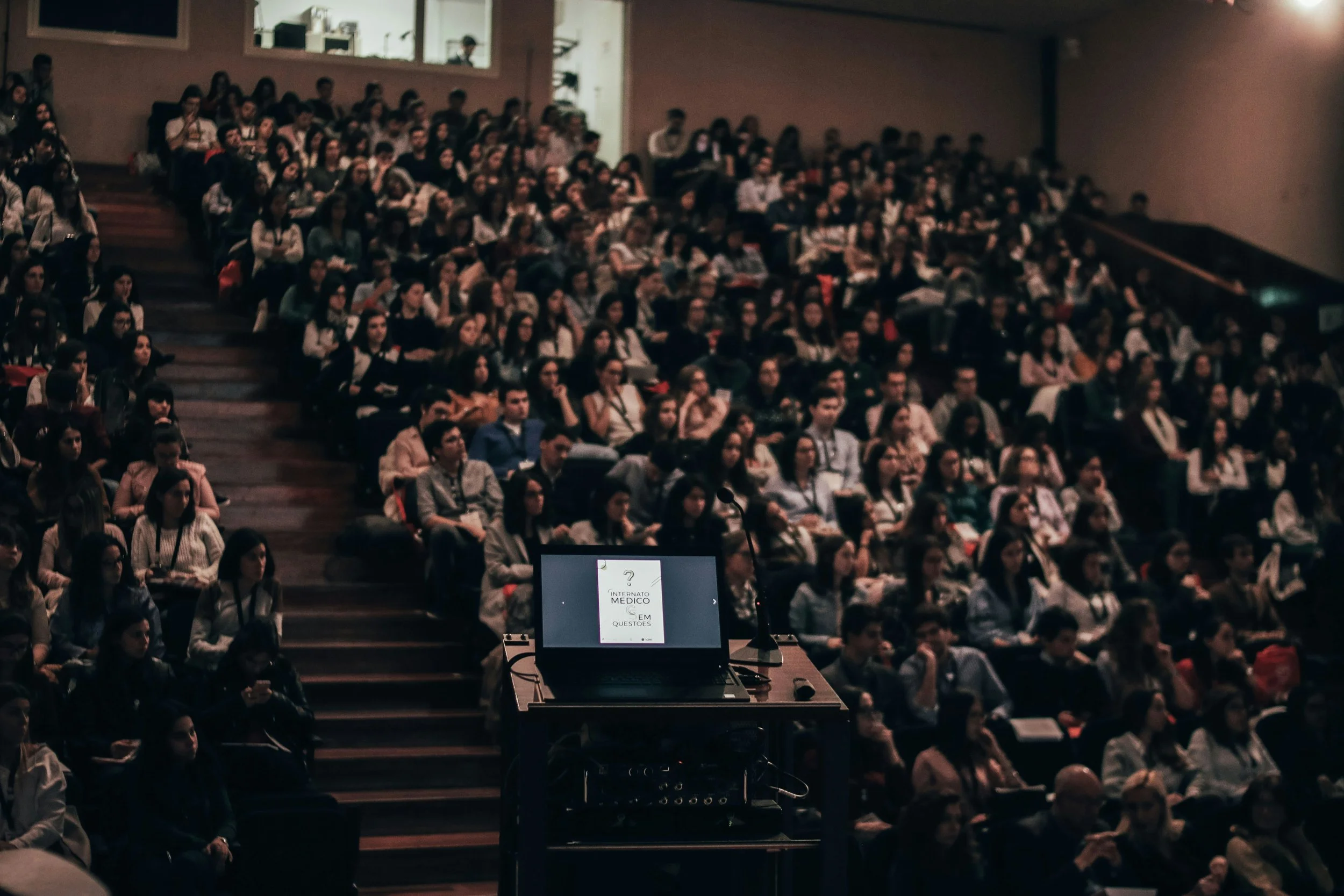 An auditorium filled with a large audience seated in rows, facing a stage with a presentation screen. The audience appears attentive, and the room is dimly lit.